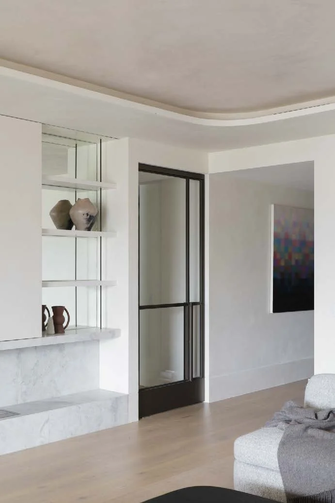 A corner of a modern living room with white walls, a ceiling with a curved molding, a black-framed glass door, and a wall-mounted art piece. There is a built-in shelf with ceramic vases and small decorative items, and part of a light-colored sofa with a blanket draped over it.