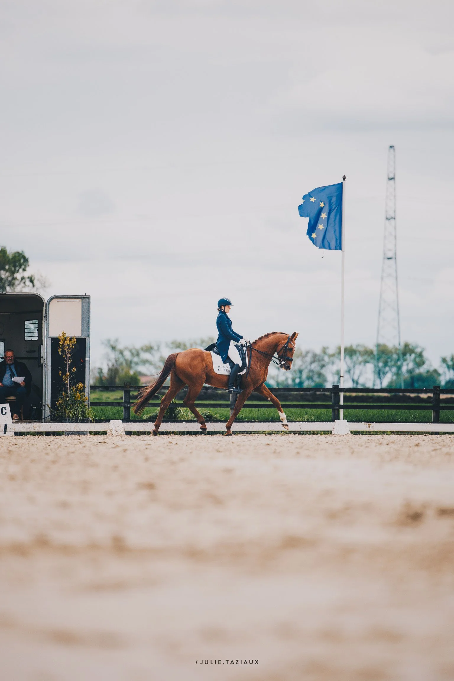 Une personne en compétition équestre, montée à cheval, portant un casque bleu, dans une enceinte de dressage avec un drapeau de l'Union européenne en arrière-plan.