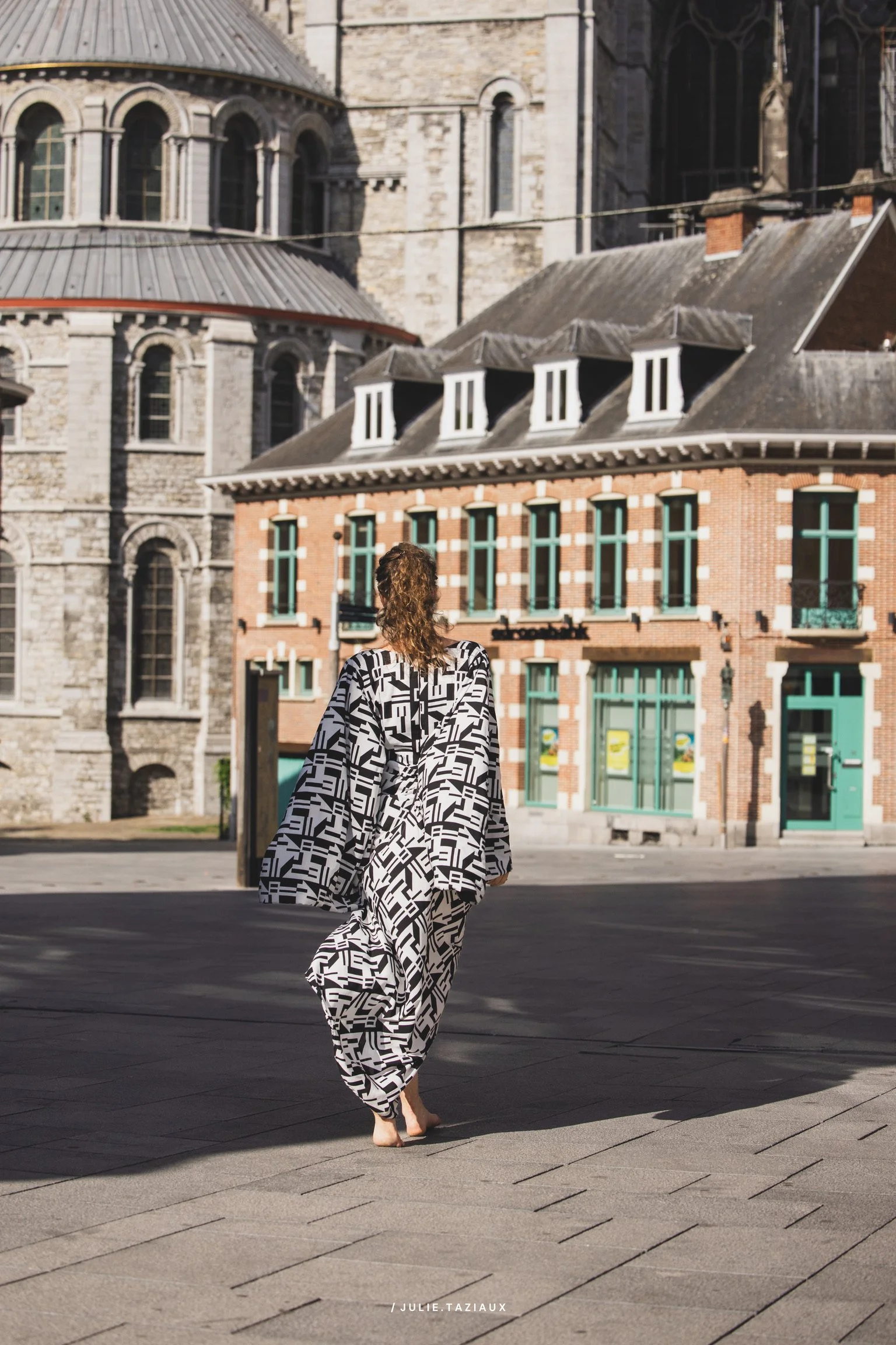Une femme marche dans une rue pavée, portant un vêtement ample à motifs géométriques noirs et blancs, avec un bâtiment ancien en pierre et une maison en briques derrière elle.