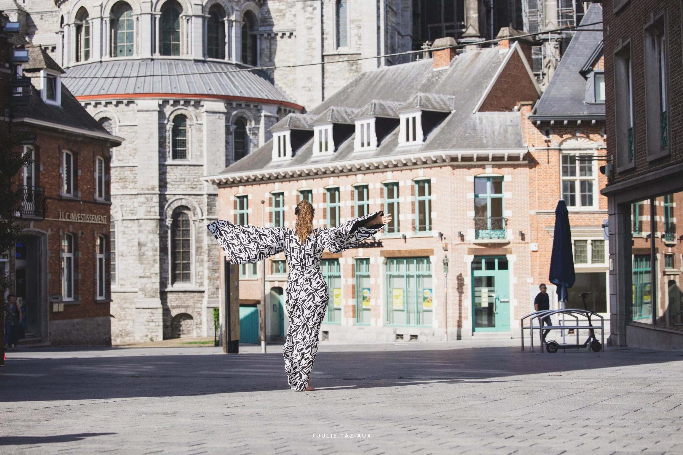 Femme en robe à motifs noirs et blancs, debout dans une place urbaine, bras ouverts, bâtiments en arrière-plan.