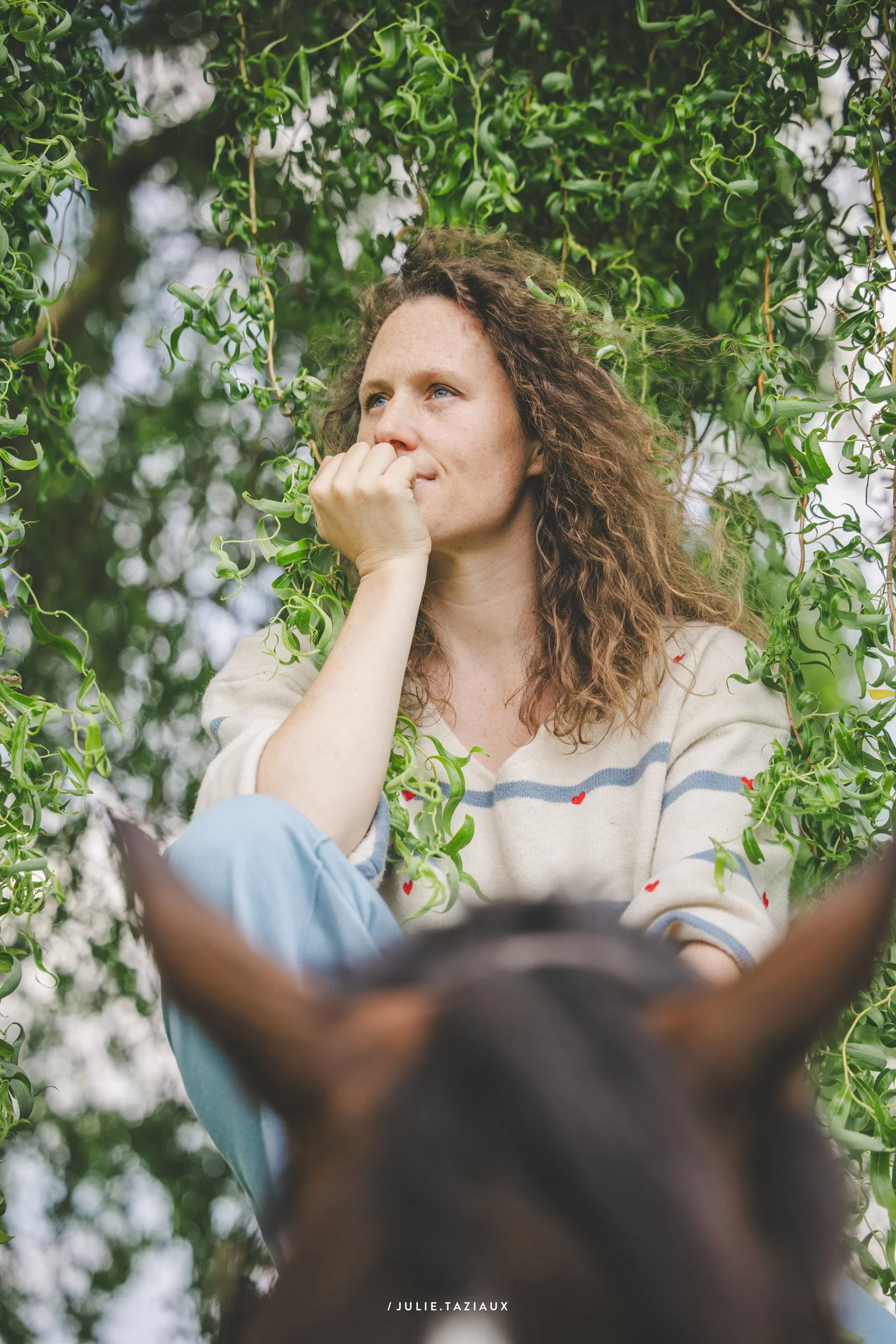 Une femme aux cheveux bouclés, assise dans un cadre verdoyant, dont seule la tête de son cheval est visible en premier plan.