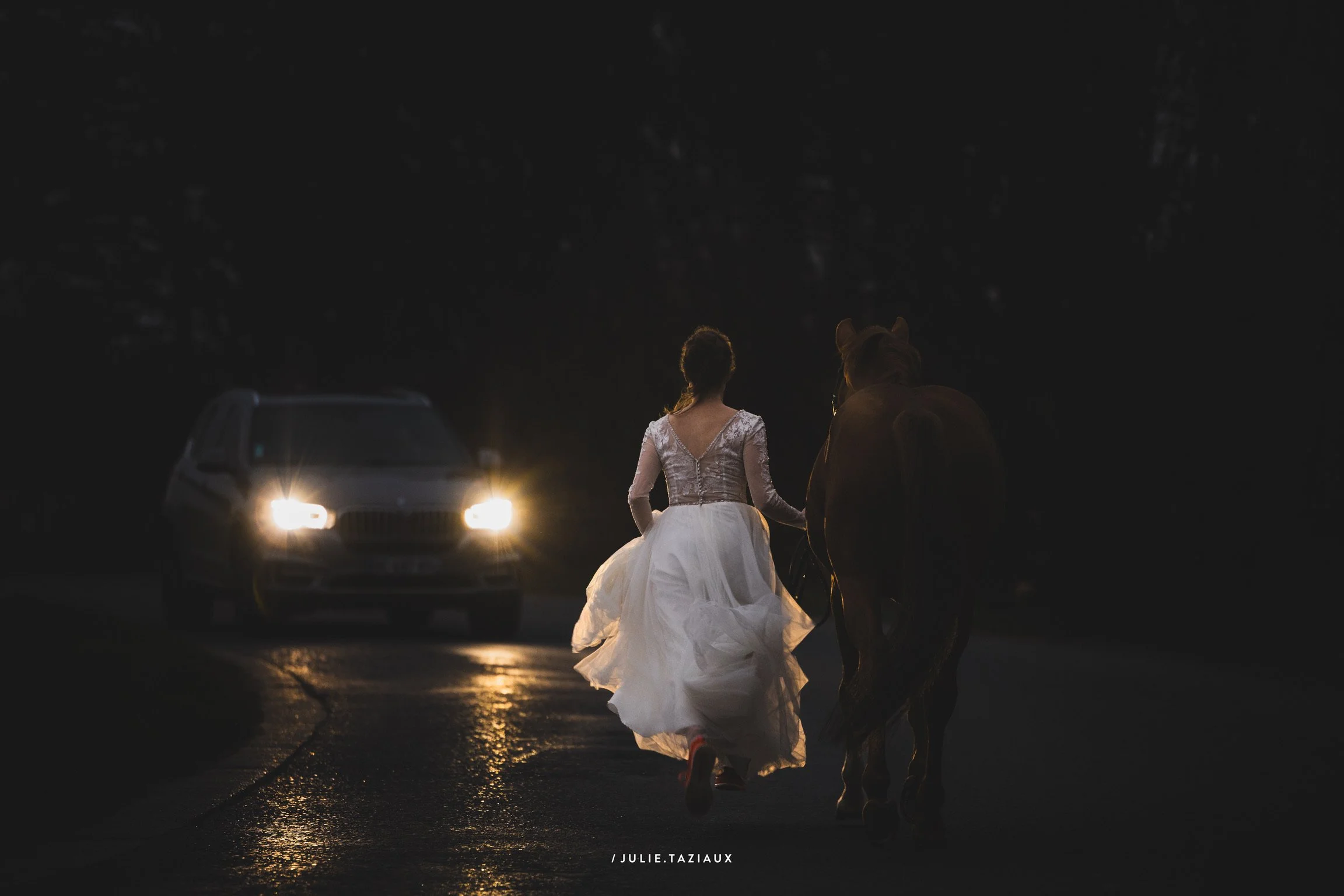 Une femme en robe de mariée marche avec un cheval sur une route sombre, avec une voiture aux phares allumés en arrière-plan.