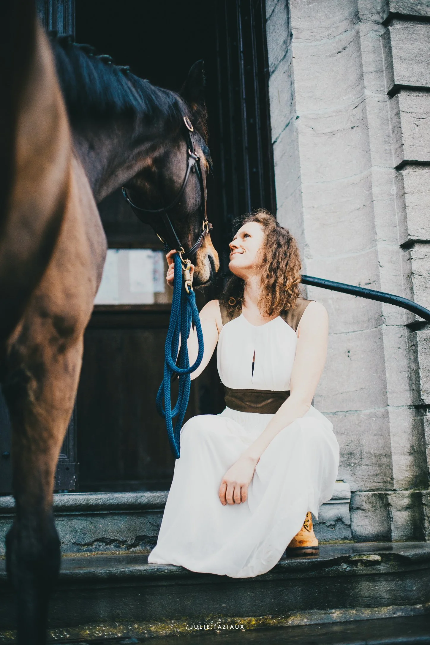 Une jeune femme en robe blanche et chaussures marron, assise sur un escalier en pierre, regarde tendrement un cheval noir avec un licol bleu, devant un bâtiment en pierre.