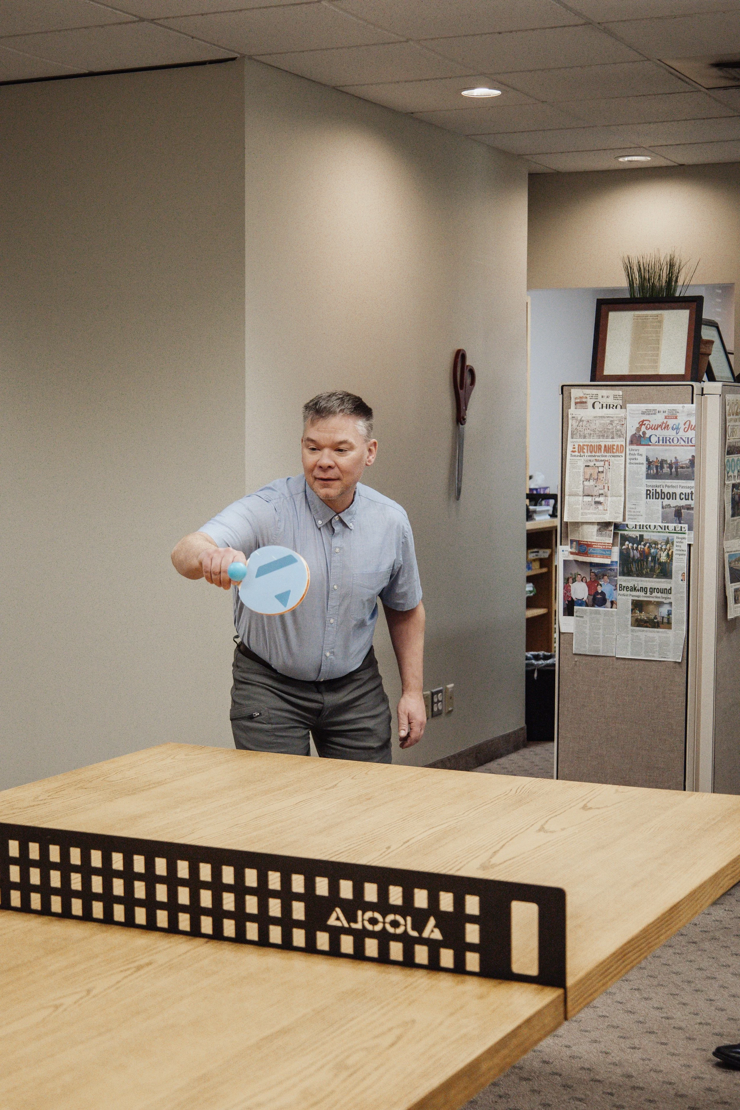 A man at Varela Engineering & Management in Spokane WA, playing ping pong indoors, holding a paddle and preparing to hit a ball, with a newspaper-covered divider in the background.