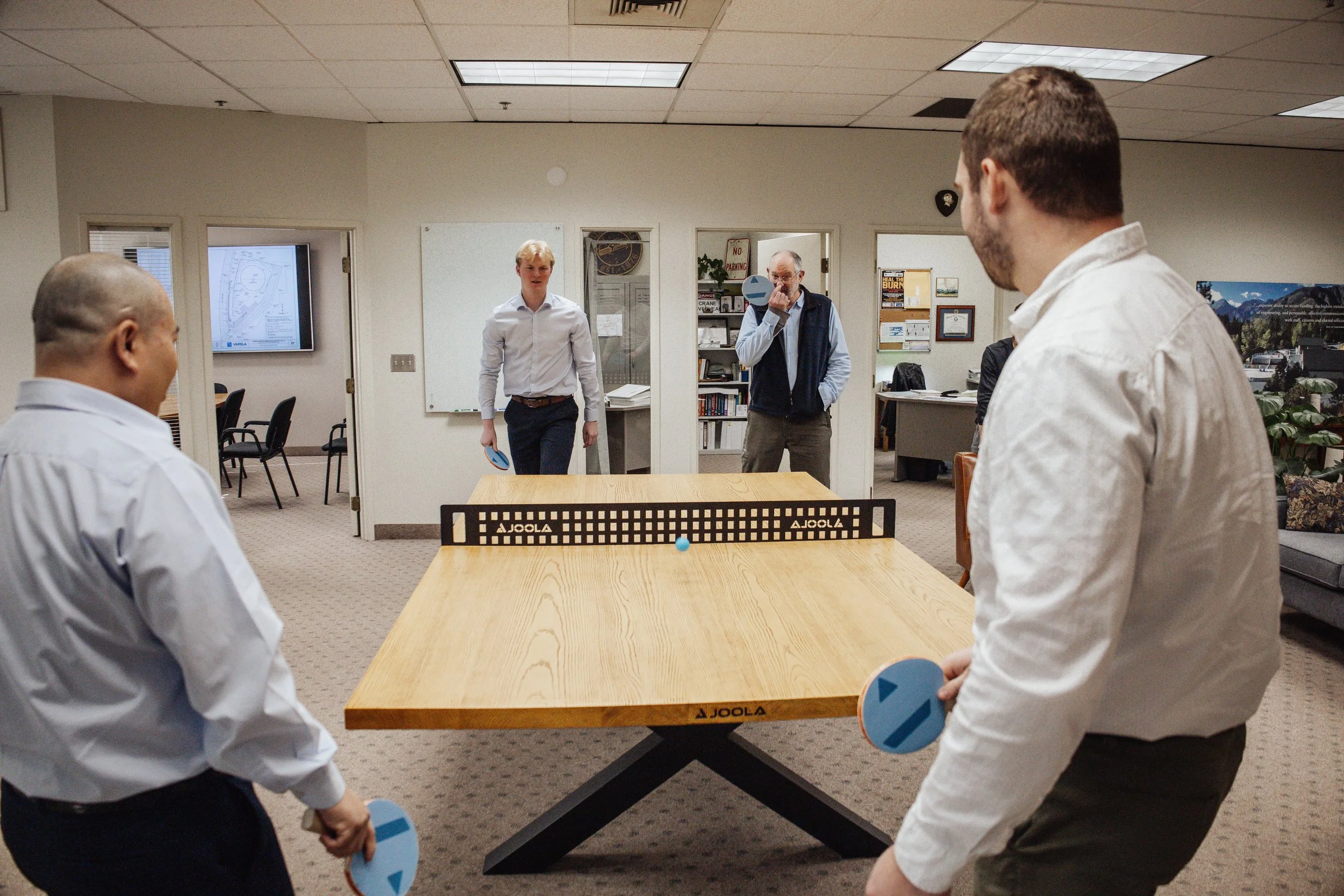 Four engineers in business attire at Varela Engineering & Management engage in company culture with a ping pong match.