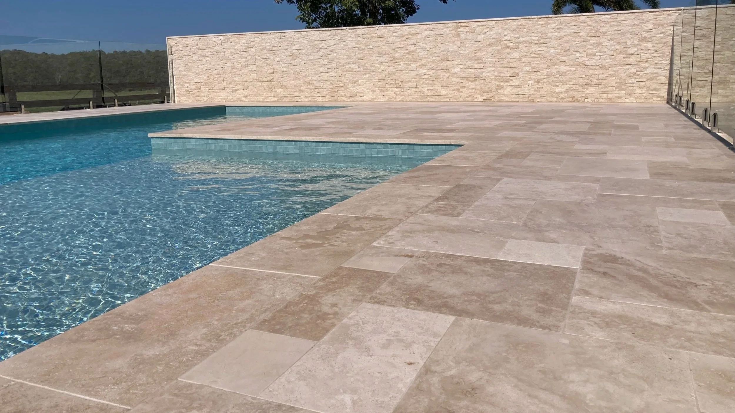 Modern outdoor pool area with clear blue water, beige stone tile flooring, a glass fence, and a beige brick wall in the background.