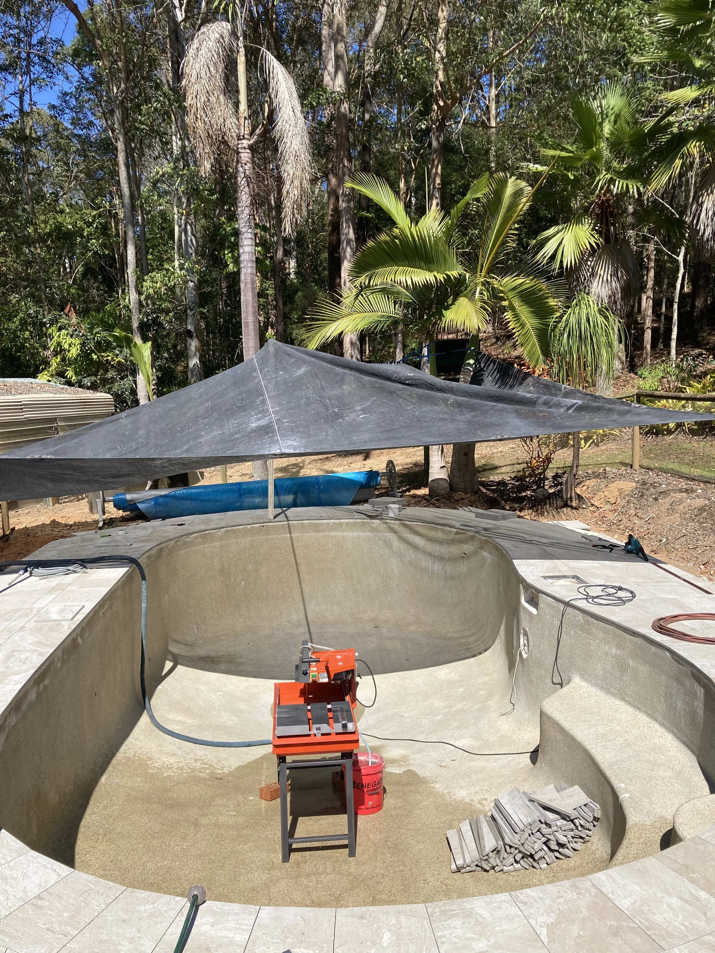 Under construction backyard swimming pool with tiles, surrounded by a patio, under a shade cloth, with trees in the background.