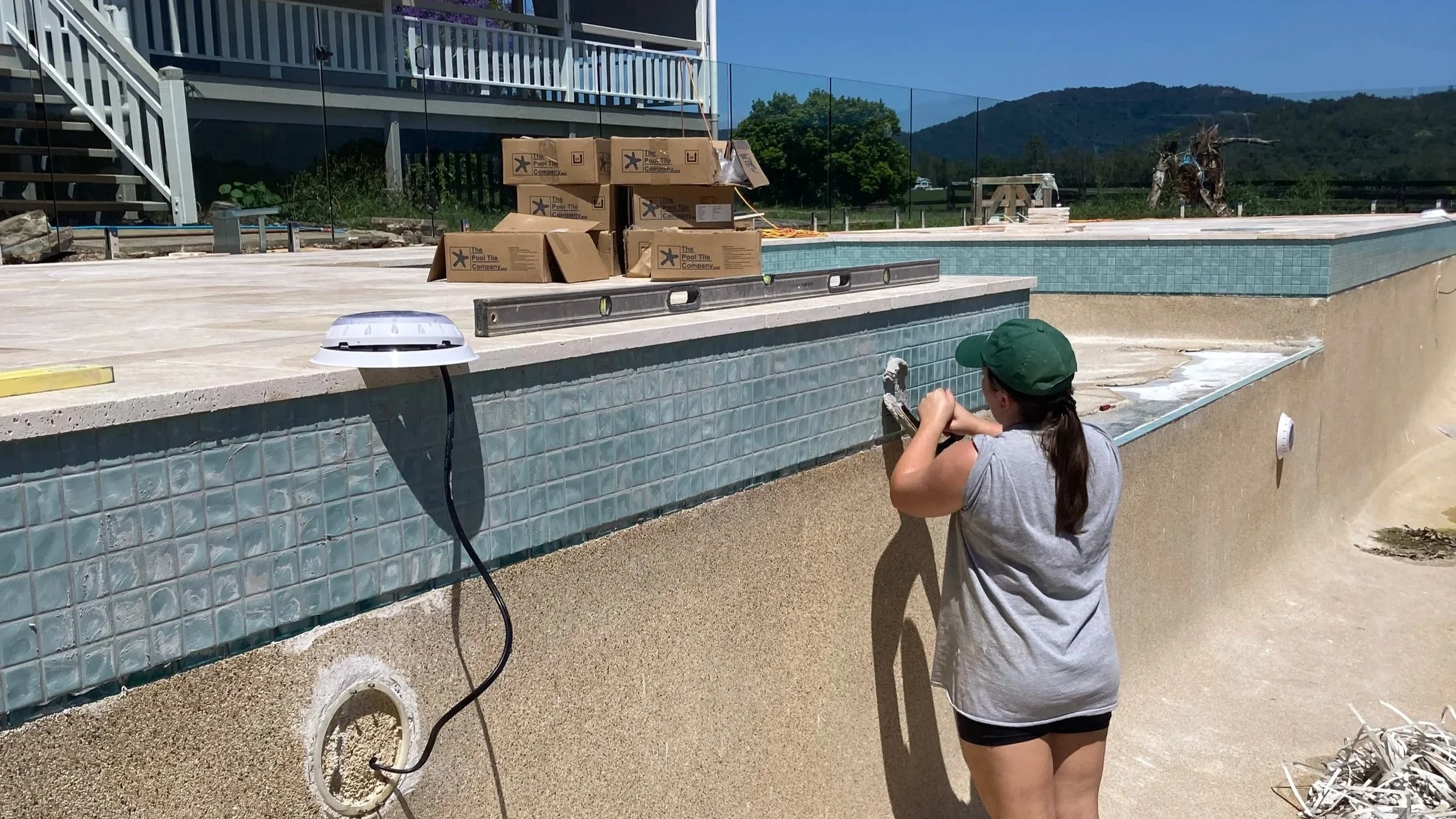 Person working on tiling the edge of an outdoor swimming pool, with construction materials and tools around.