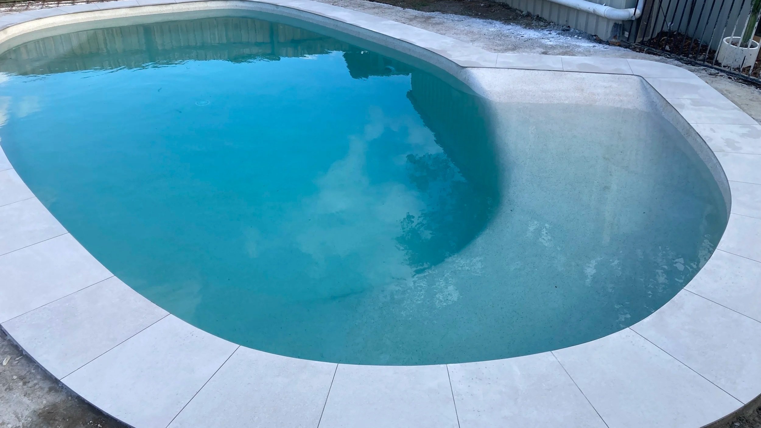 An empty in-ground swimming pool with light-colored tile surround, partially filled with water reflecting the sky and nearby trees, with unfinished yard area around it.