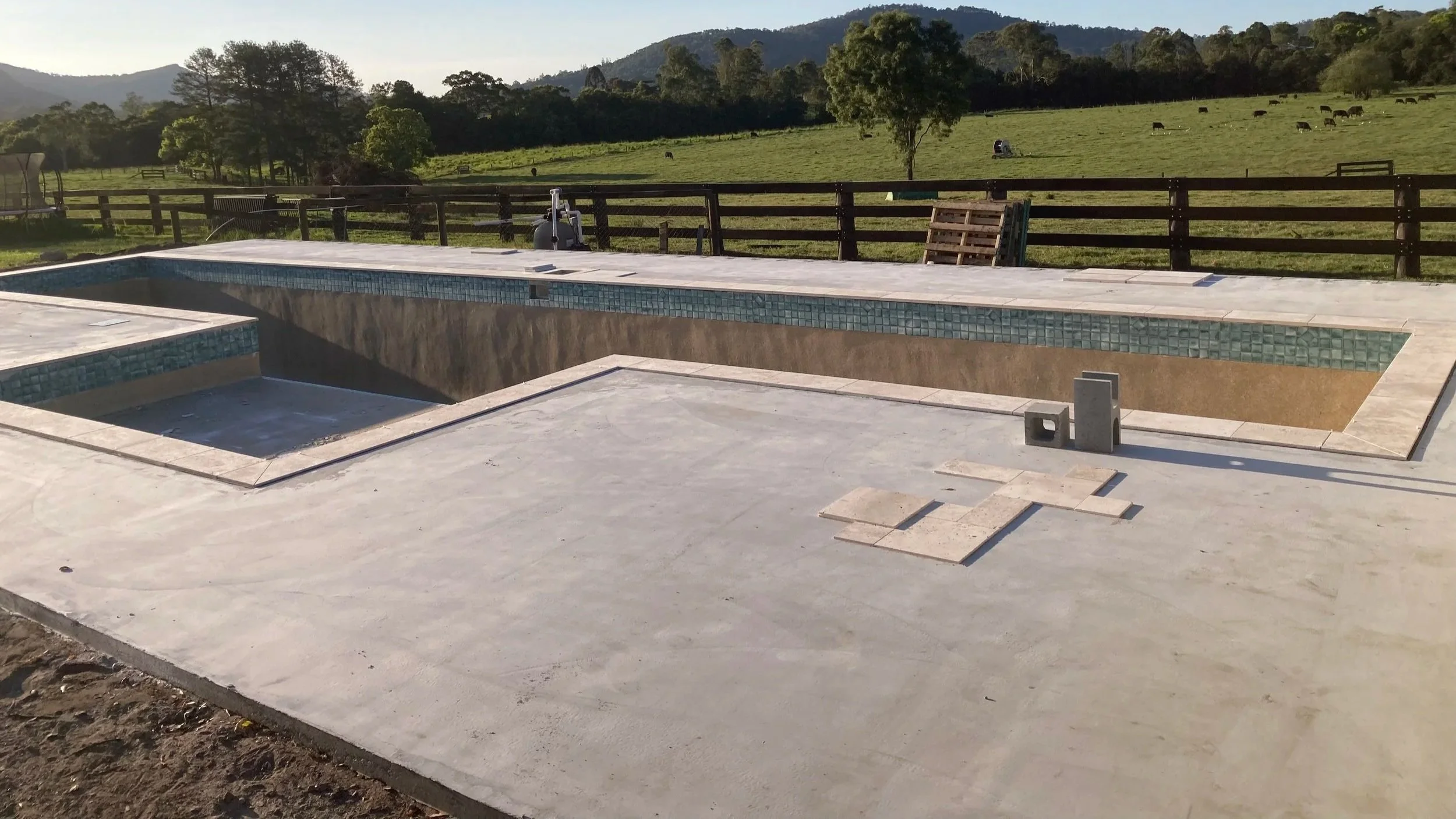 View of an unfinished swimming pool with a tiled interior, surrounded by a concrete patio, overlooking a rural landscape with green fields and cows grazing.