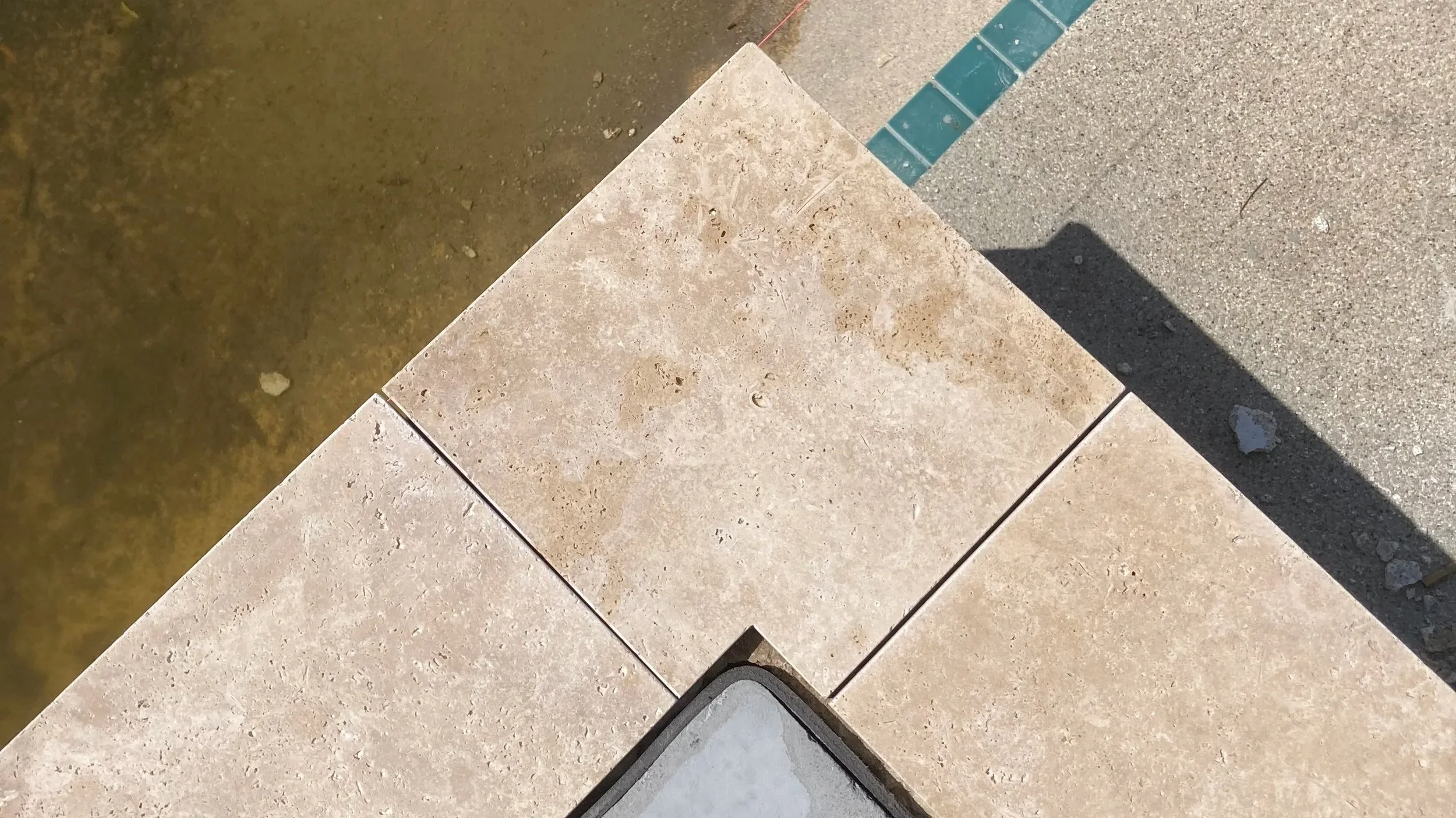 Close-up of beige stone tiles on a corner with a gray utility cover and a cast shadow on the surface.