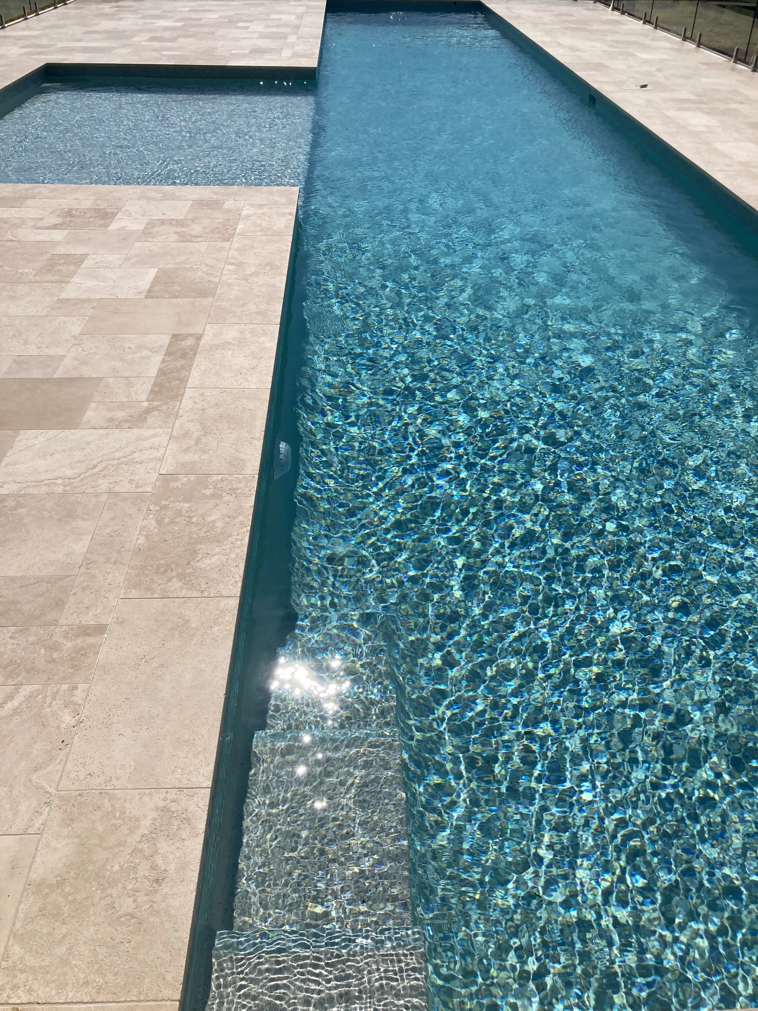 A swimming pool with clear blue water and a small shallow section, surrounded by beige tiled decking.