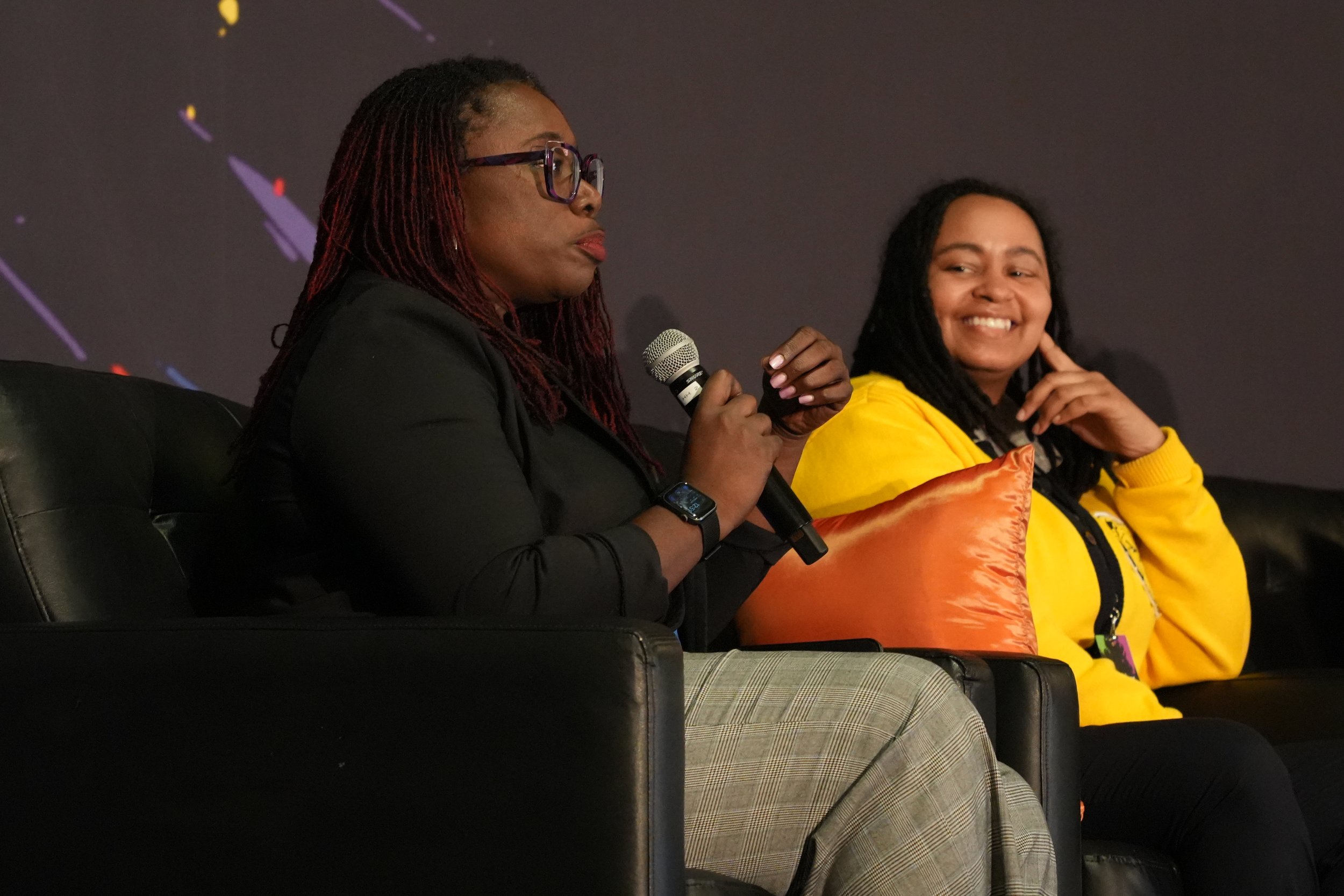 Two women sitting on black leather chairs. The woman on the left is speaking into a microphone, wearing glasses, a black blazer, and a watch. The woman on the right is smiling, wearing a yellow hoodie and has long dreadlocks.