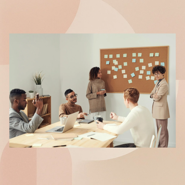 A diverse group of five young adults in a modern office meeting room, engaged in a discussion around a wooden table with laptops and notebooks. One person is presenting in front of a bulletin board with sticky notes, while others listen and participate.