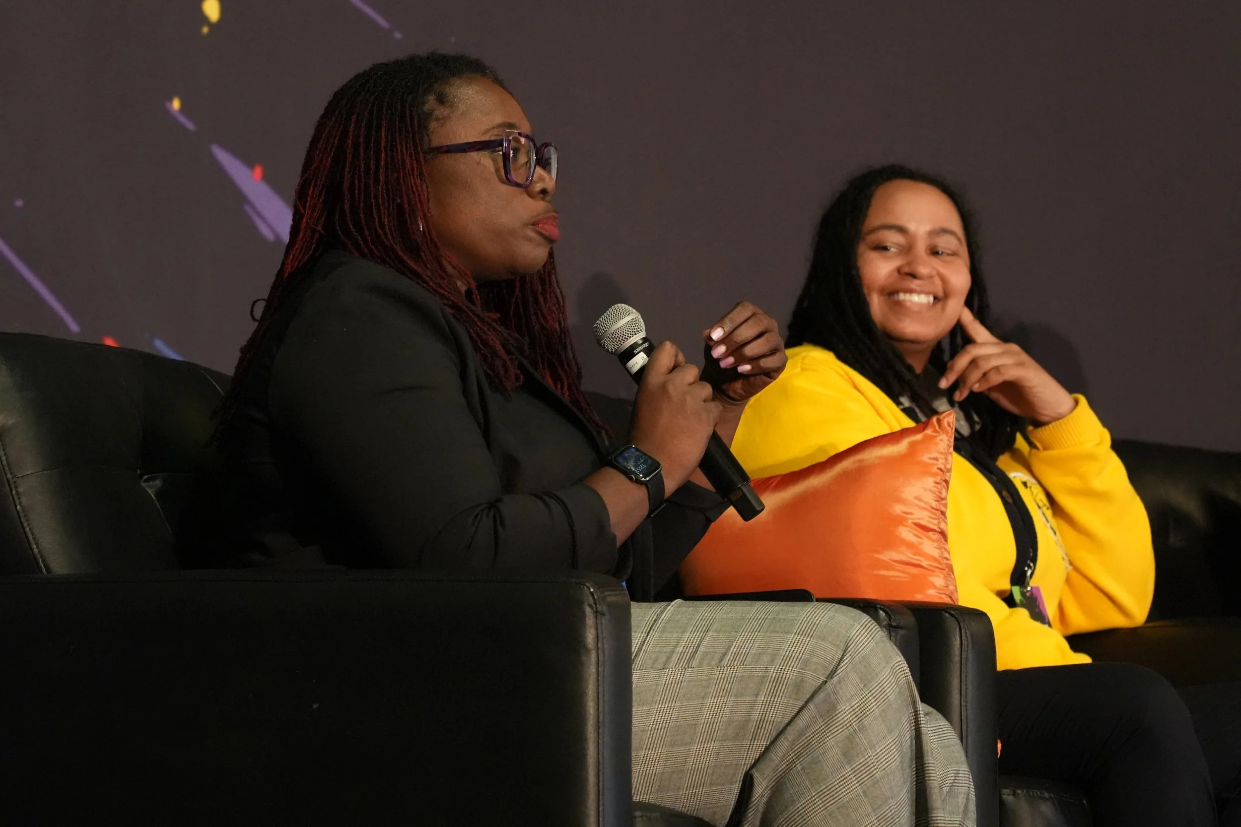Two women sitting on a stage, one holding a microphone and the other smiling, with a dark background.