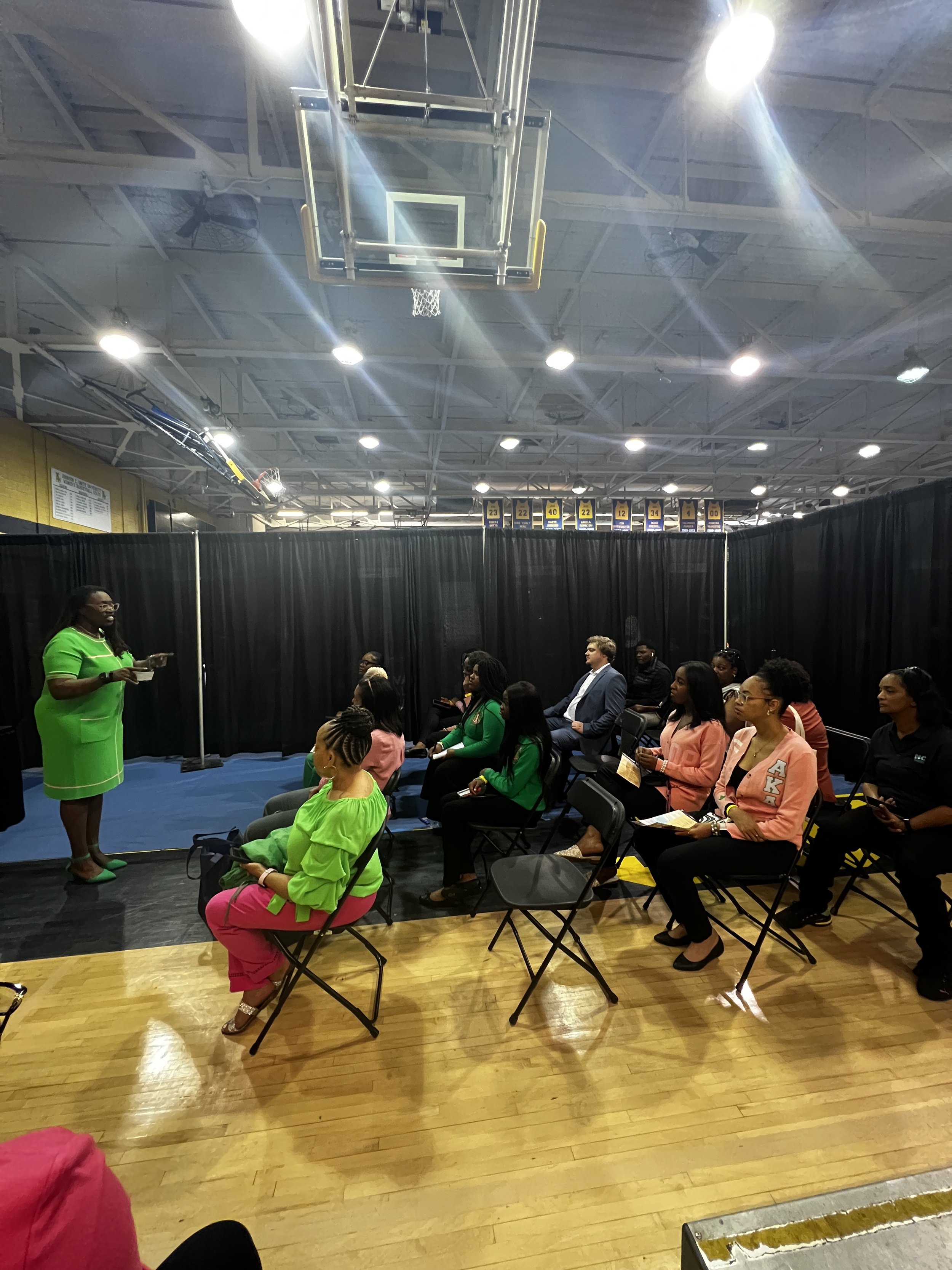 A woman in a bright green dress and matching shoes is standing in front of an audience in a gymnasium, giving a presentation or speech. The audience sits on black chairs, with some dressed in colorful attire, and black curtains separate the presentation area from the rest of the gym.
