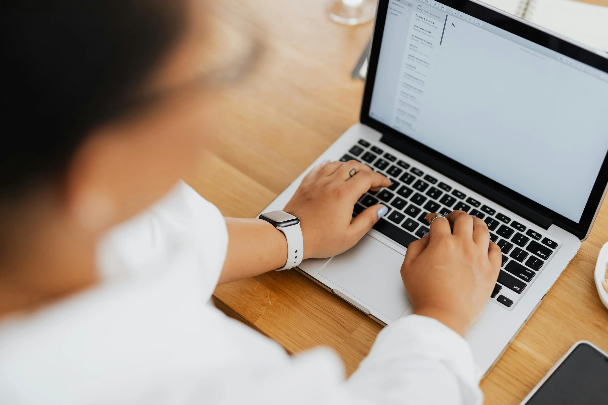 Person working on a laptop at a wooden desk, wearing a white watch and light blue nail polish.