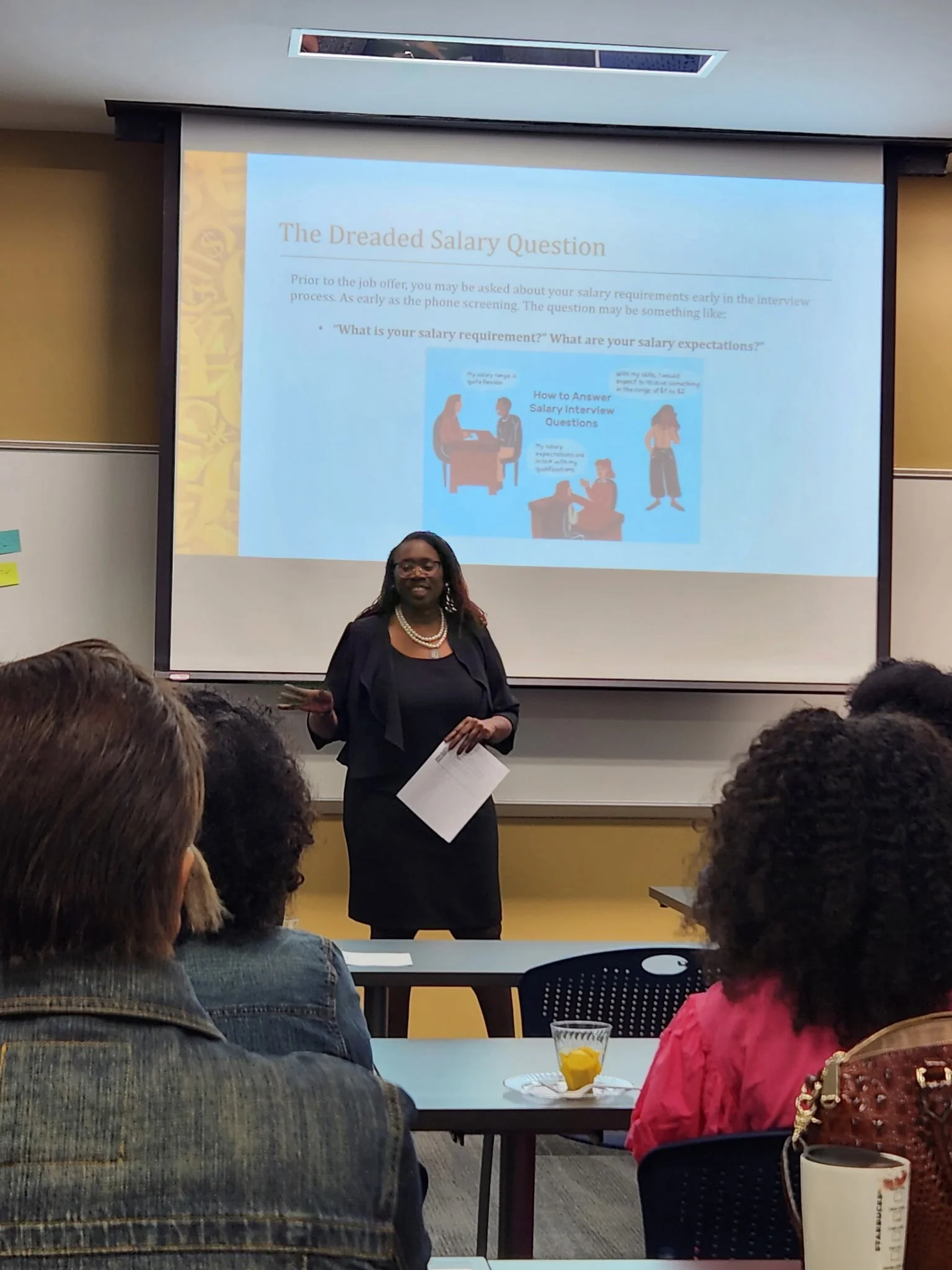 A woman giving a presentation in a classroom, with a slide titled 'The Dreaded Salary Question' projected behind her. Several audience members are seated and listening, with one having a drink on the table.