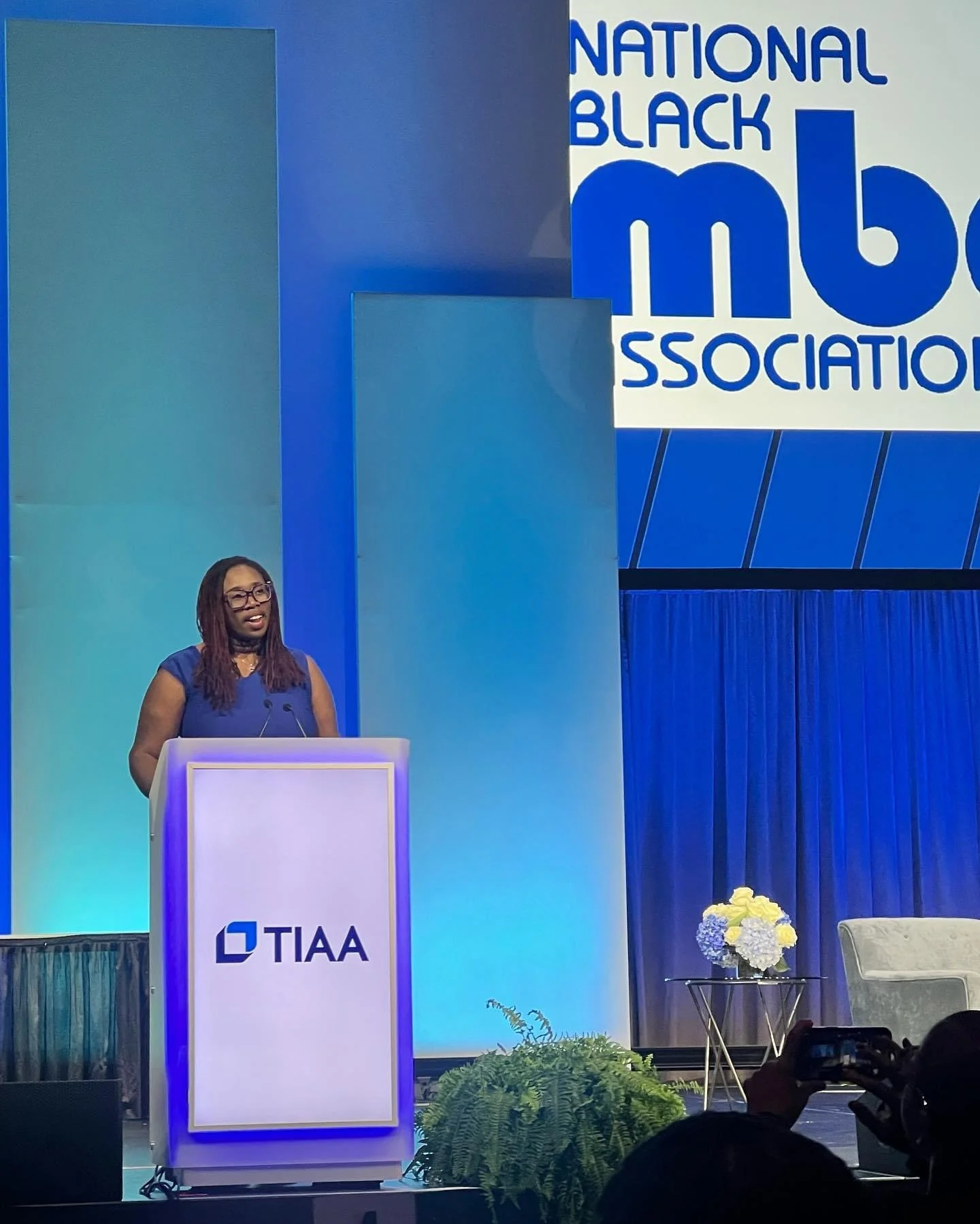 A woman with glasses and dreadlocks speaking at a podium during a conference for the National Black MBA Association, with a large blue and white background and floral arrangement on stage.