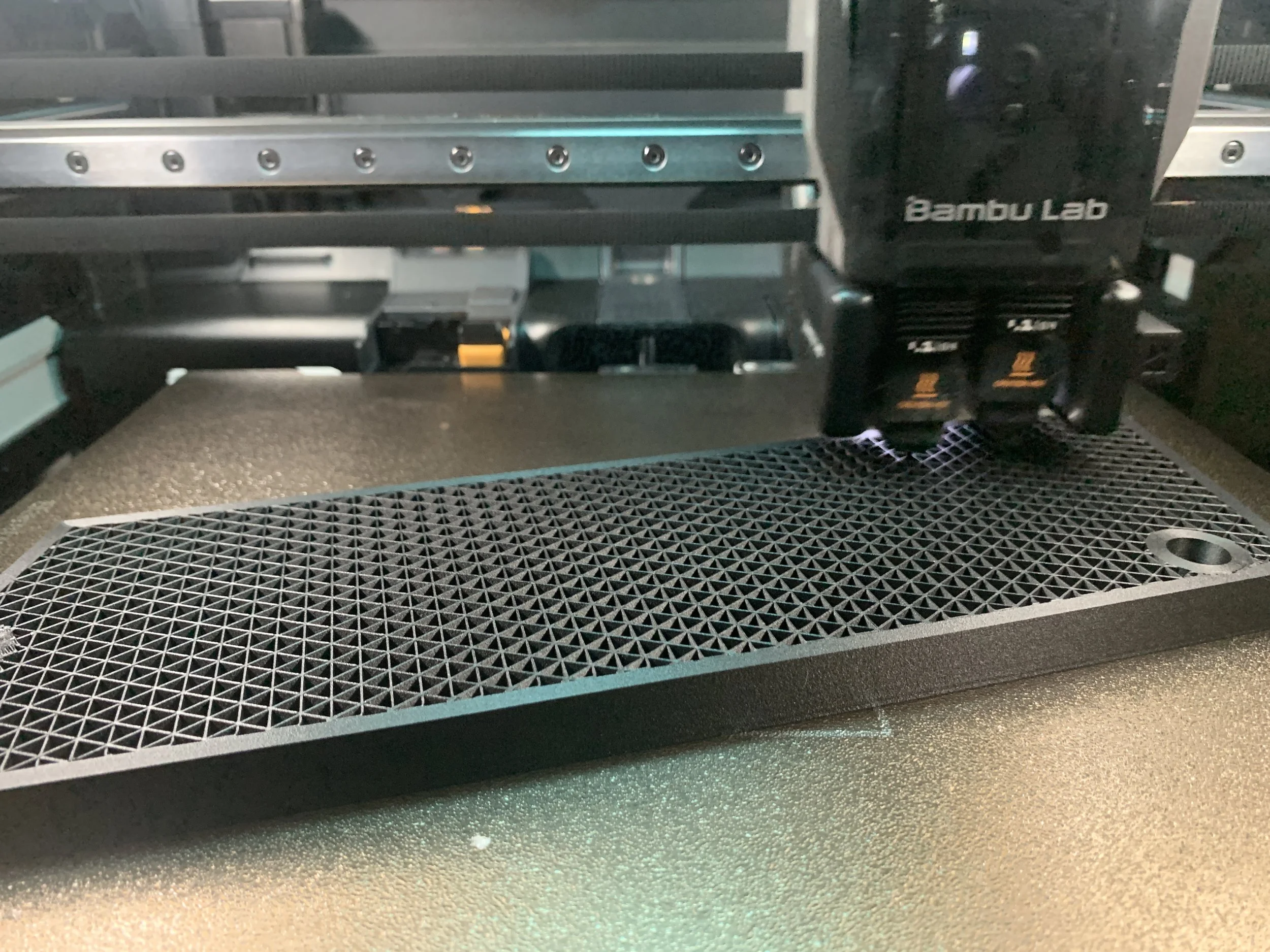 Close-up of a 3D printer's print bed and extruder, labeled 'Bambu Lab,' in a manufacturing workshop, printing in triangular infill.