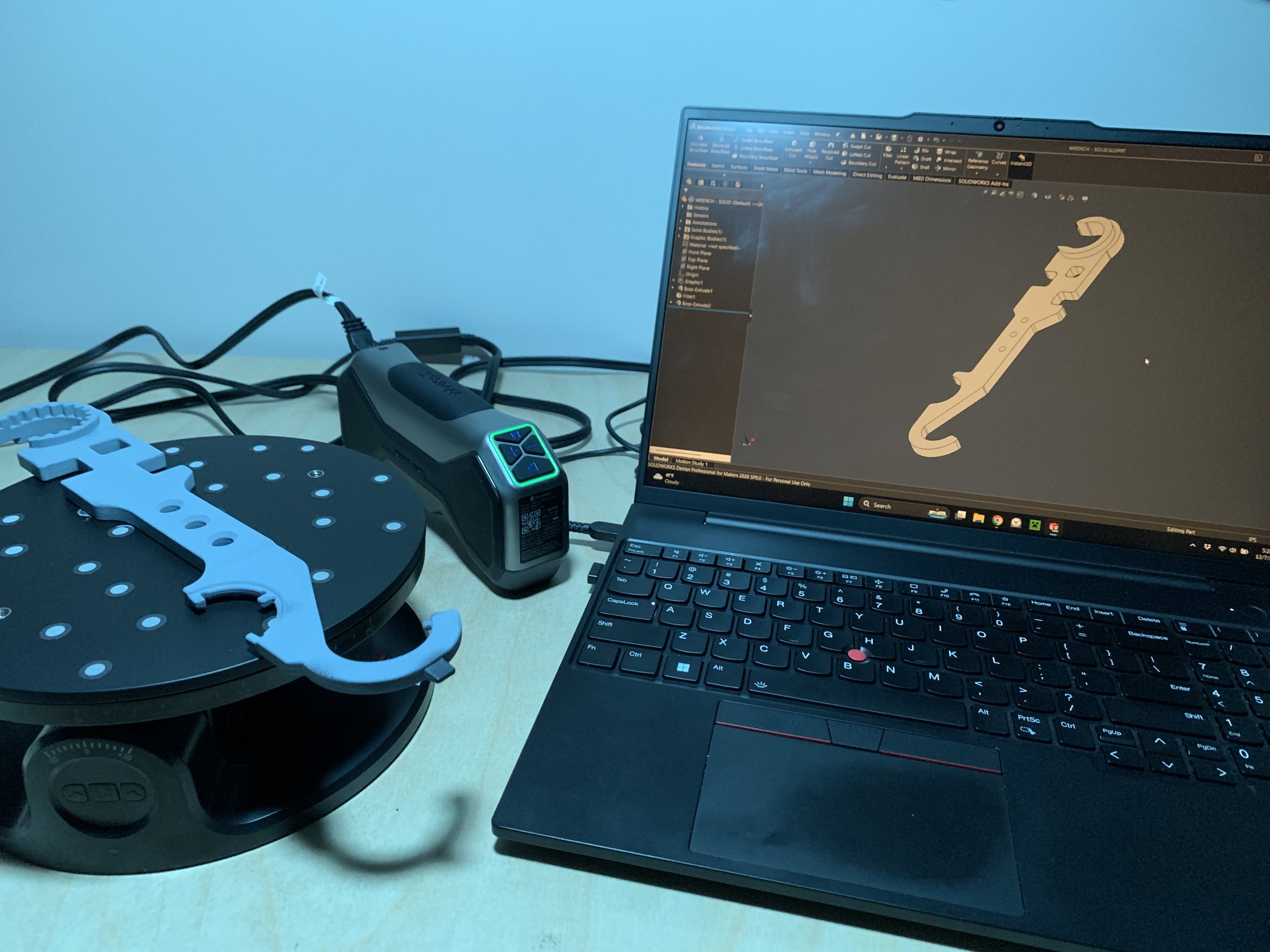 Laptop displaying a CAD design of a mechanical and a circular scanning platform with a mechanical part on it connected to a power source on a desk.
