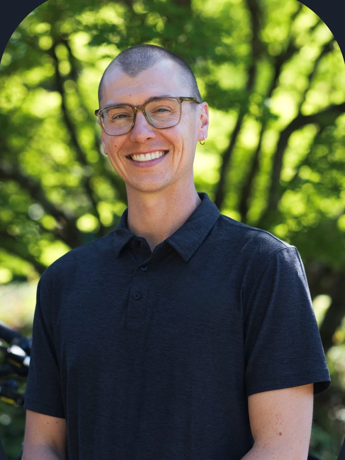 Smiling person wearing glasses and a dark polo shirt outdoors in a sunlit forest