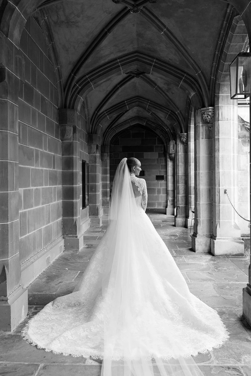 A bride in a wedding dress with a veil standing under gothic arches in a stone corridor, looking to the side. Bridal Hair and Makeup by makeup artist merton muaremi