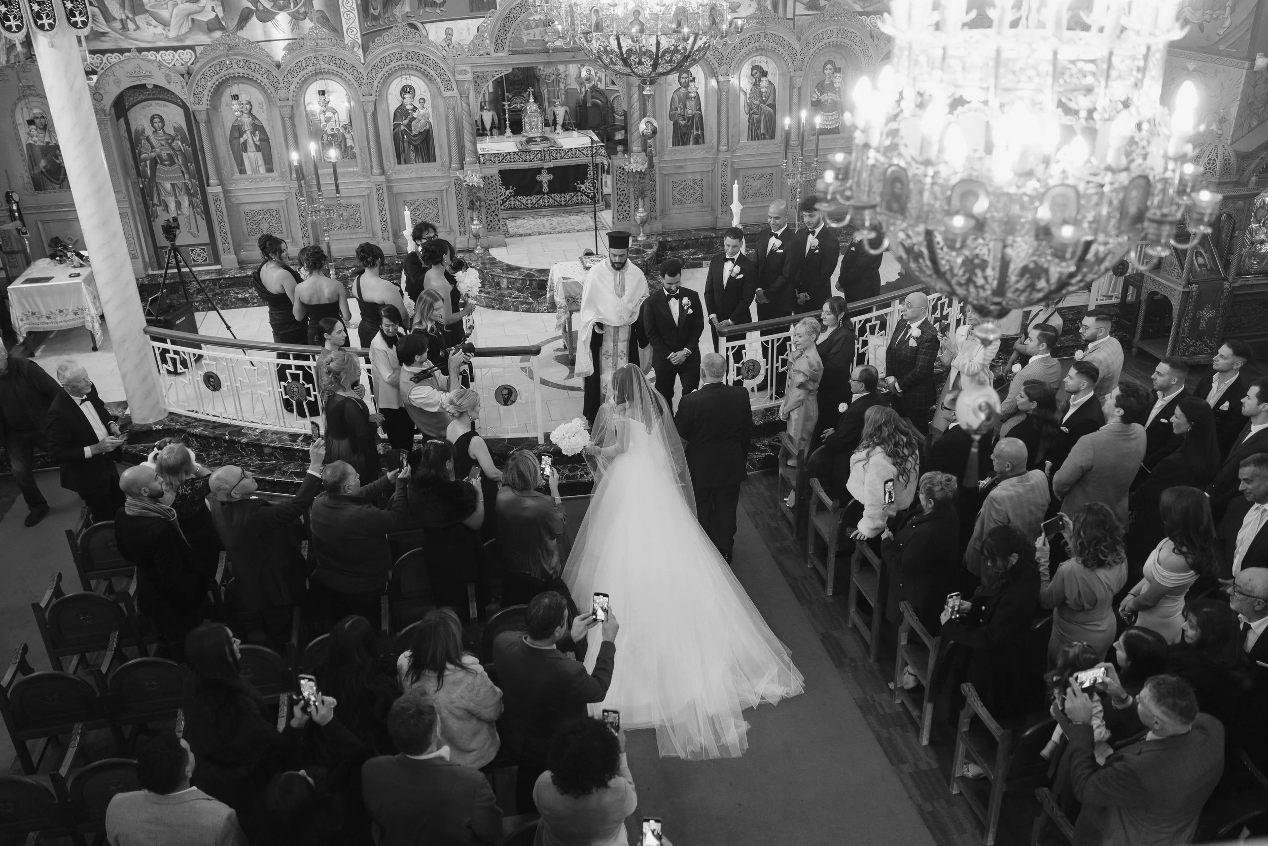 A wedding ceremony inside a church with the bride and groom at the altar, surrounded by guests and officiant, with religious icons and statues on the wall.
