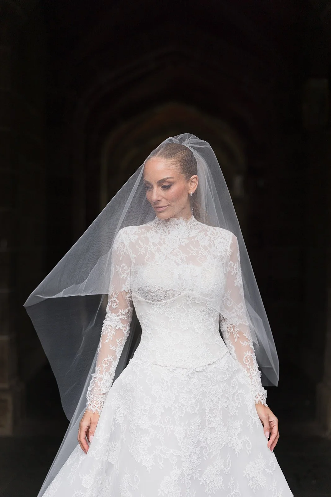 A bride in a white lace wedding dress with long sleeves and high neckline, wearing a veil that drapes over her face and shoulders, standing in a dimly lit archway. Bridal Hair and Makeup by makeup artist merton muaremi