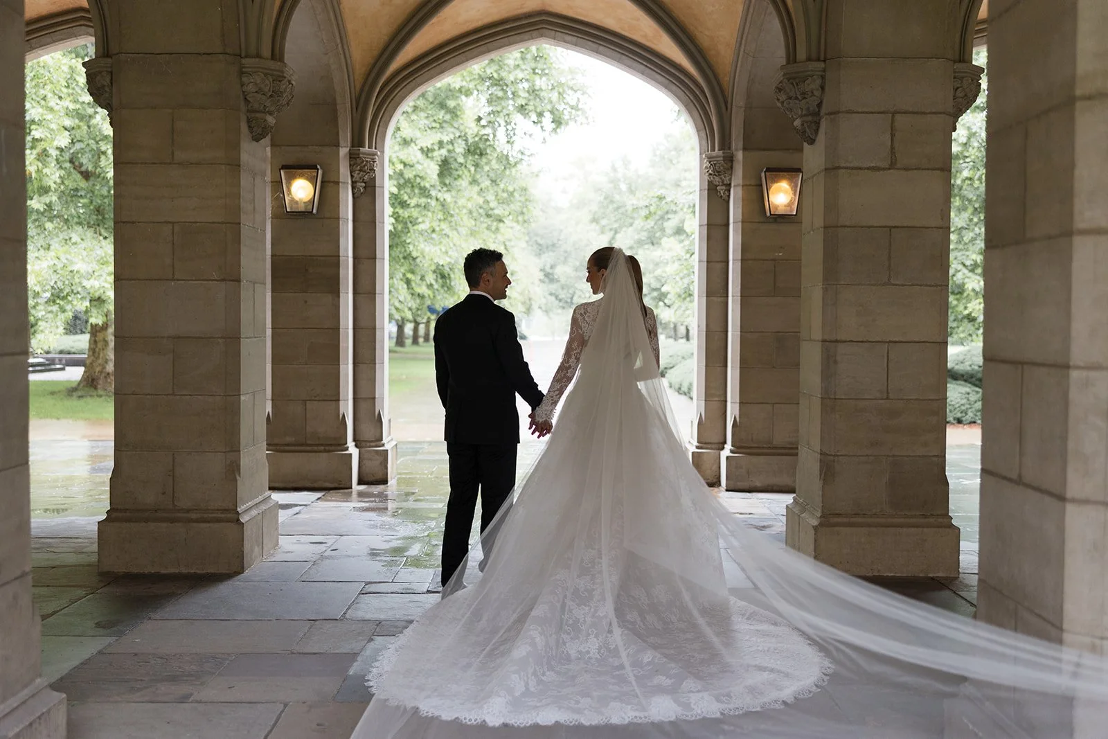 Bride and groom holding hands under stone archway, with trees in the background, on their wedding day. Bridal Hair and Makeup by makeup artist merton muaremi