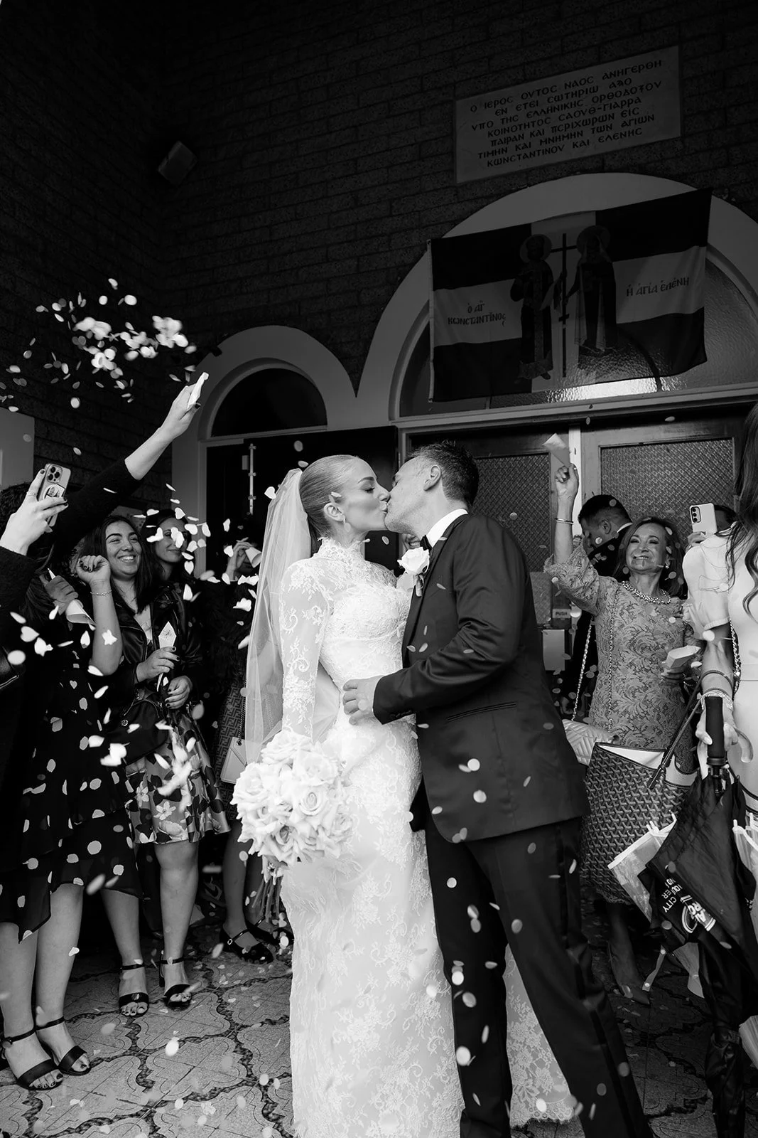 A bride and groom kiss in front of celebrating guests throwing confetti at their wedding ceremony.