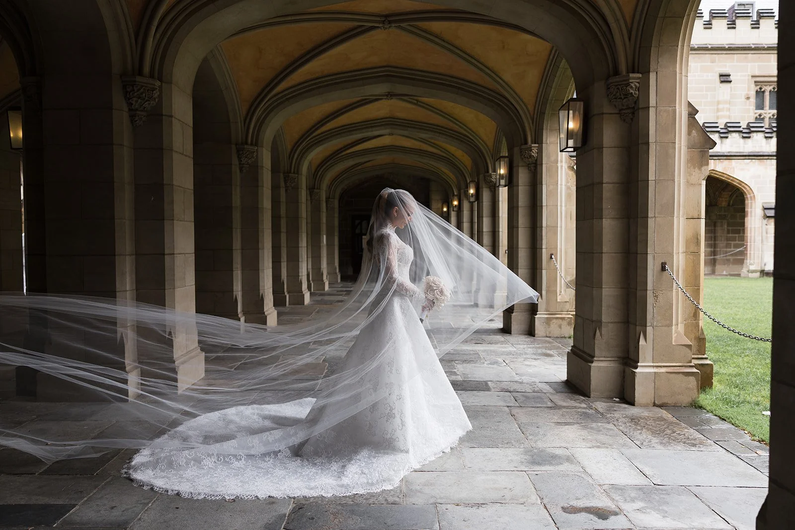 Bridal Hair and Makeup by makeup artist merton muaremi. A bride in a white wedding gown, holding a bouquet, standing under a stone archway with a long veil flowing behind her, in a historic outdoor walkway with stone columns and a grassy area. 