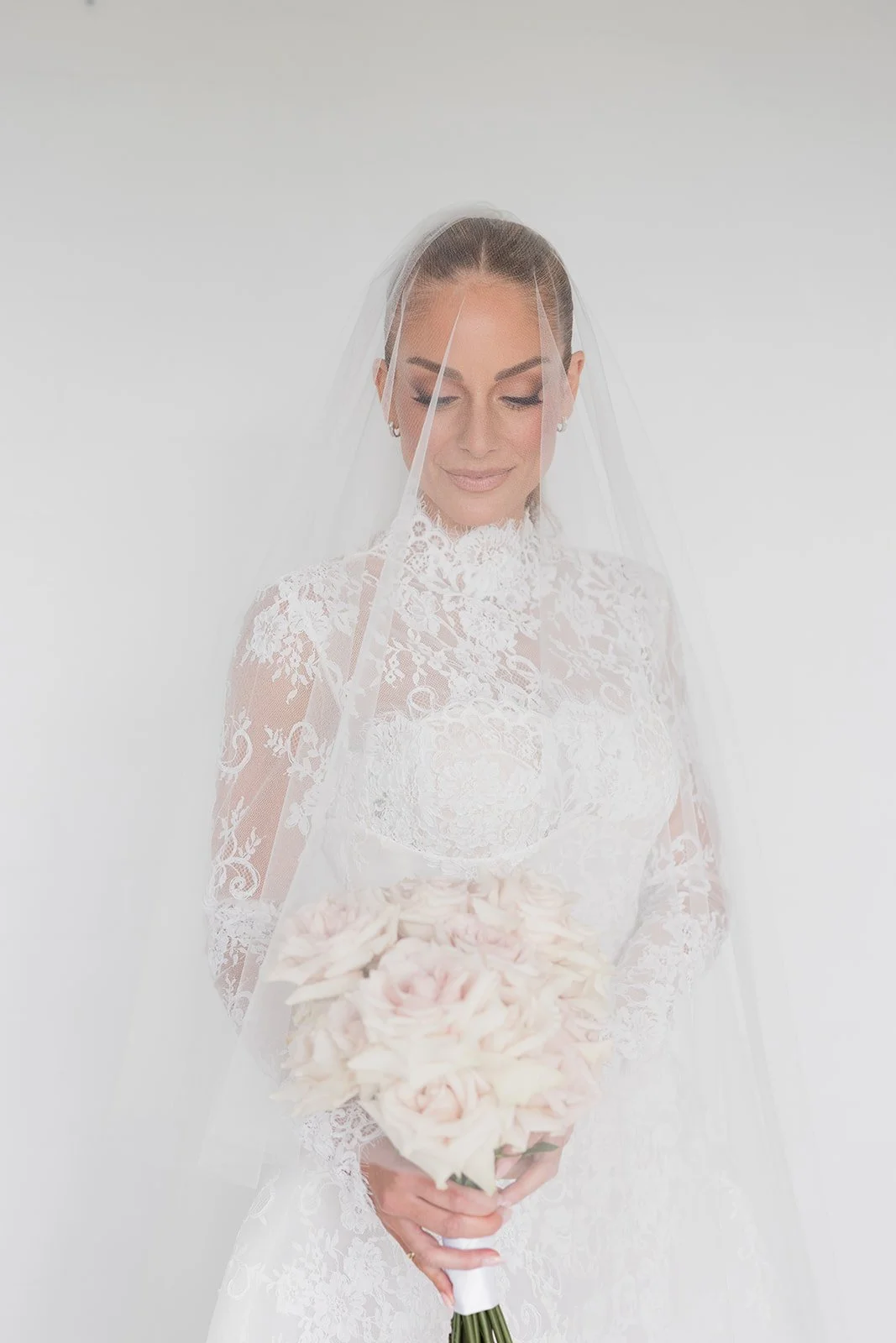 A bride in a white lace wedding dress with a veil, holding a bouquet of light pink and white roses, standing against a plain light background. Bridal Hair and Makeup by makeup artist merton muaremi