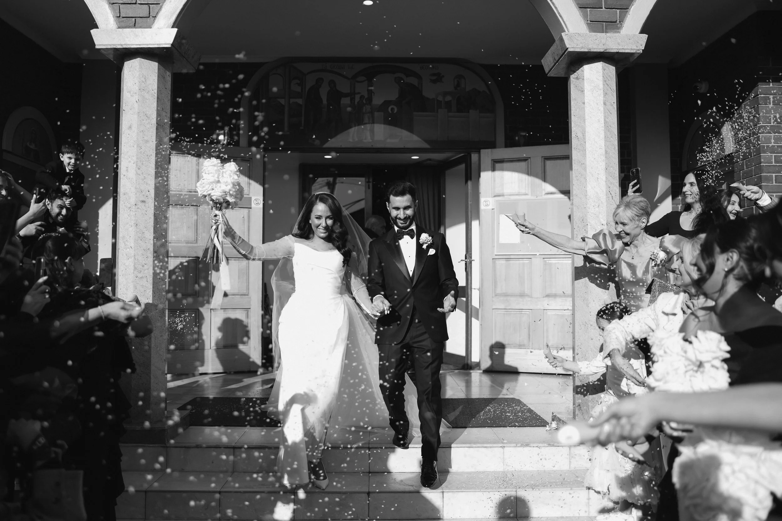 Black and white photograph of a bride and groom exiting a building, smiling, surrounded by guests throwing confetti.