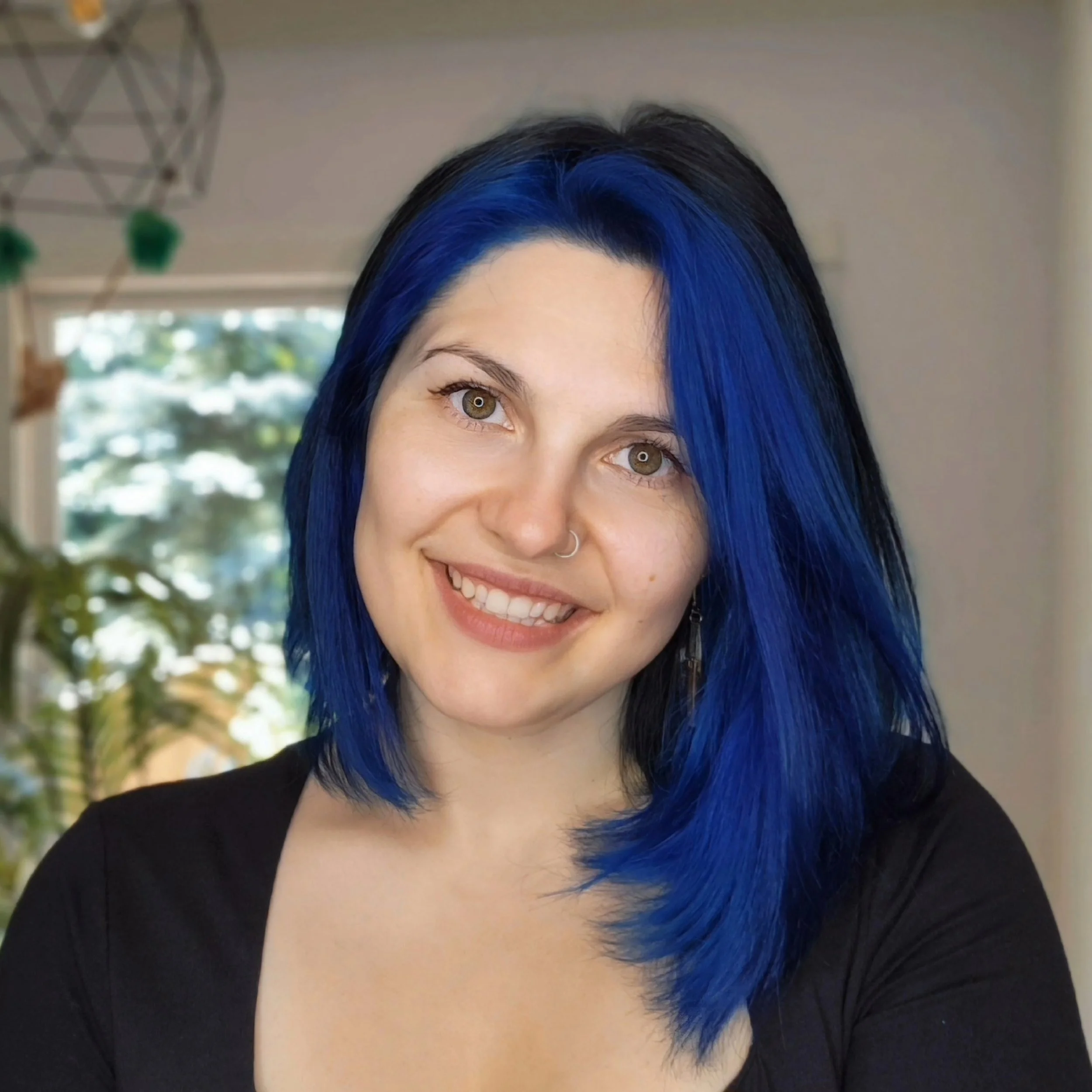 A woman with bright blue hair, smiling, with a neutral background and plants near a window.