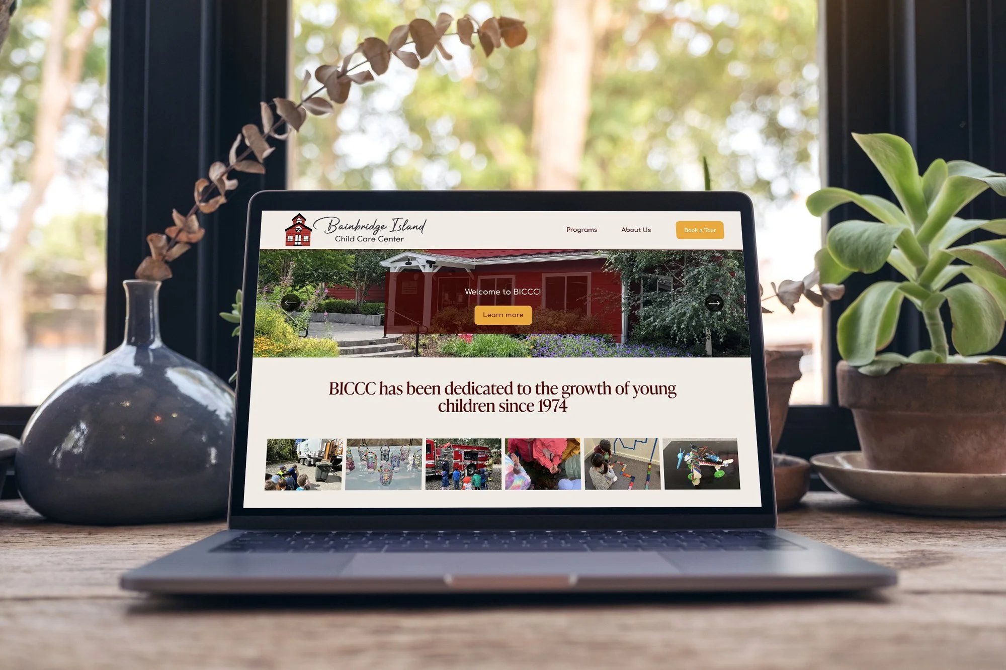Laptop on a wooden table displaying the homepage of Bainbridge Island Child Care Center website, with potted plants and a window showing greenery in the background.