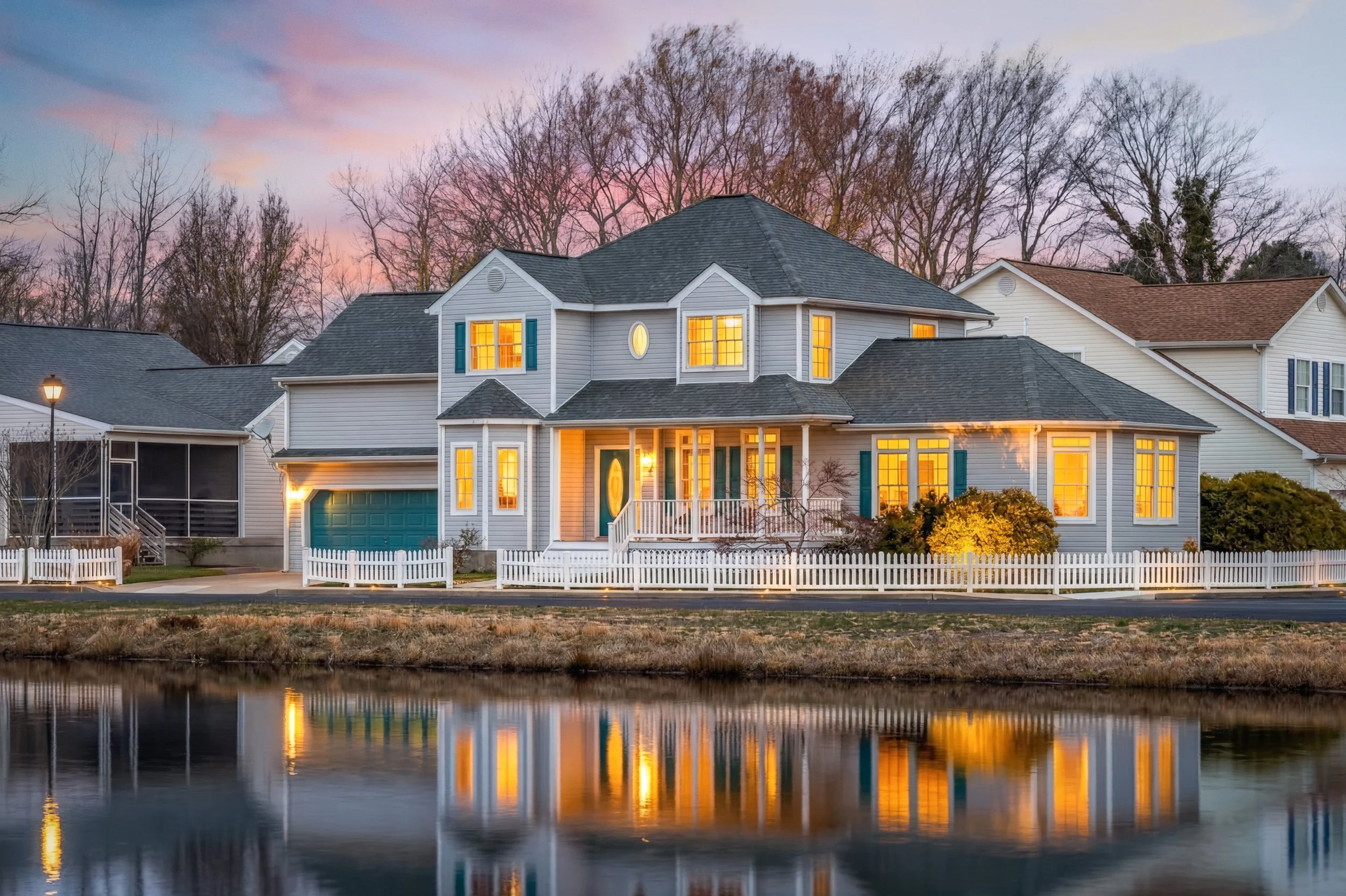 A large, two-story house with a reflecting pond in front, illuminated with warm interior lights during twilight, with trees in the background and a white picket fence around the yard.
