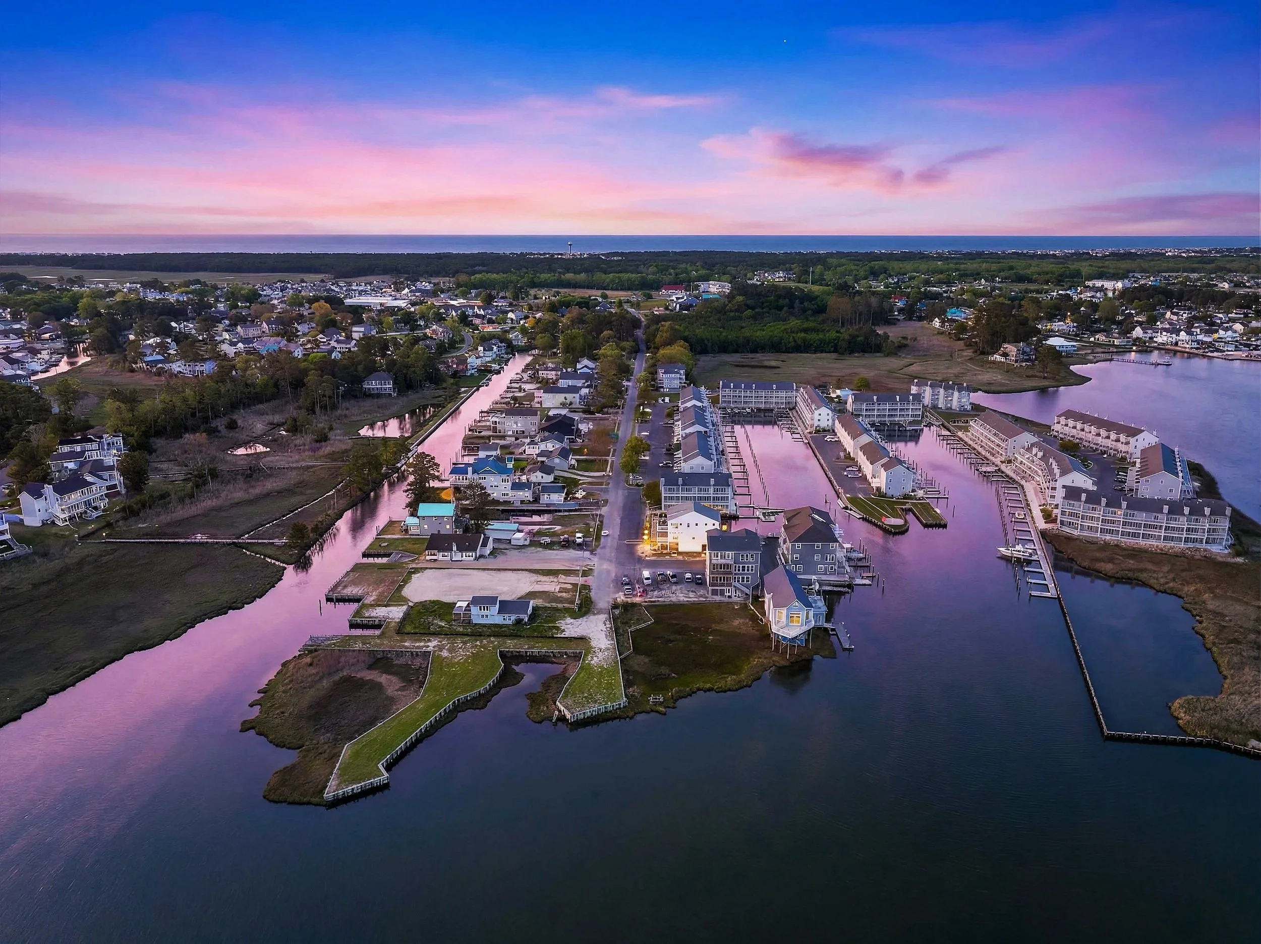 Aerial view of a suburban neighborhood at sunset, with houses along water channels, green spaces, and a large body of water on the horizon under a colorful sky.