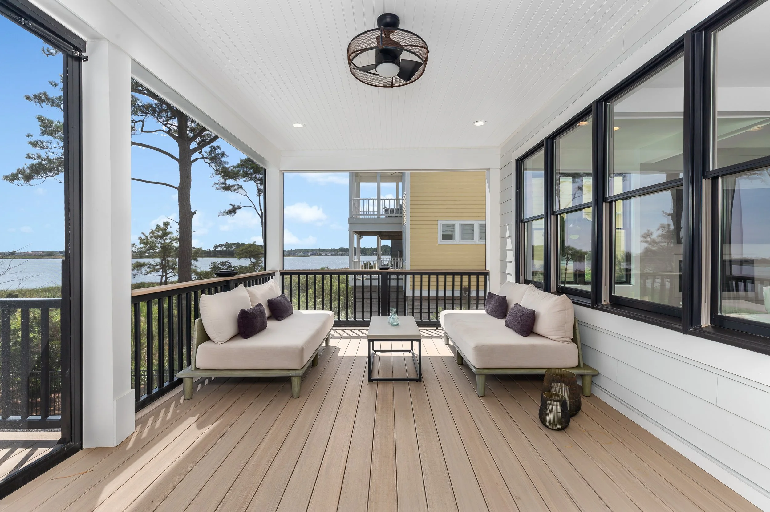 Balcony with white sofas, black pillows, and a small table overlooking a water scene with trees and a house in the distance, enclosed by a black railing and sliding glass doors.
