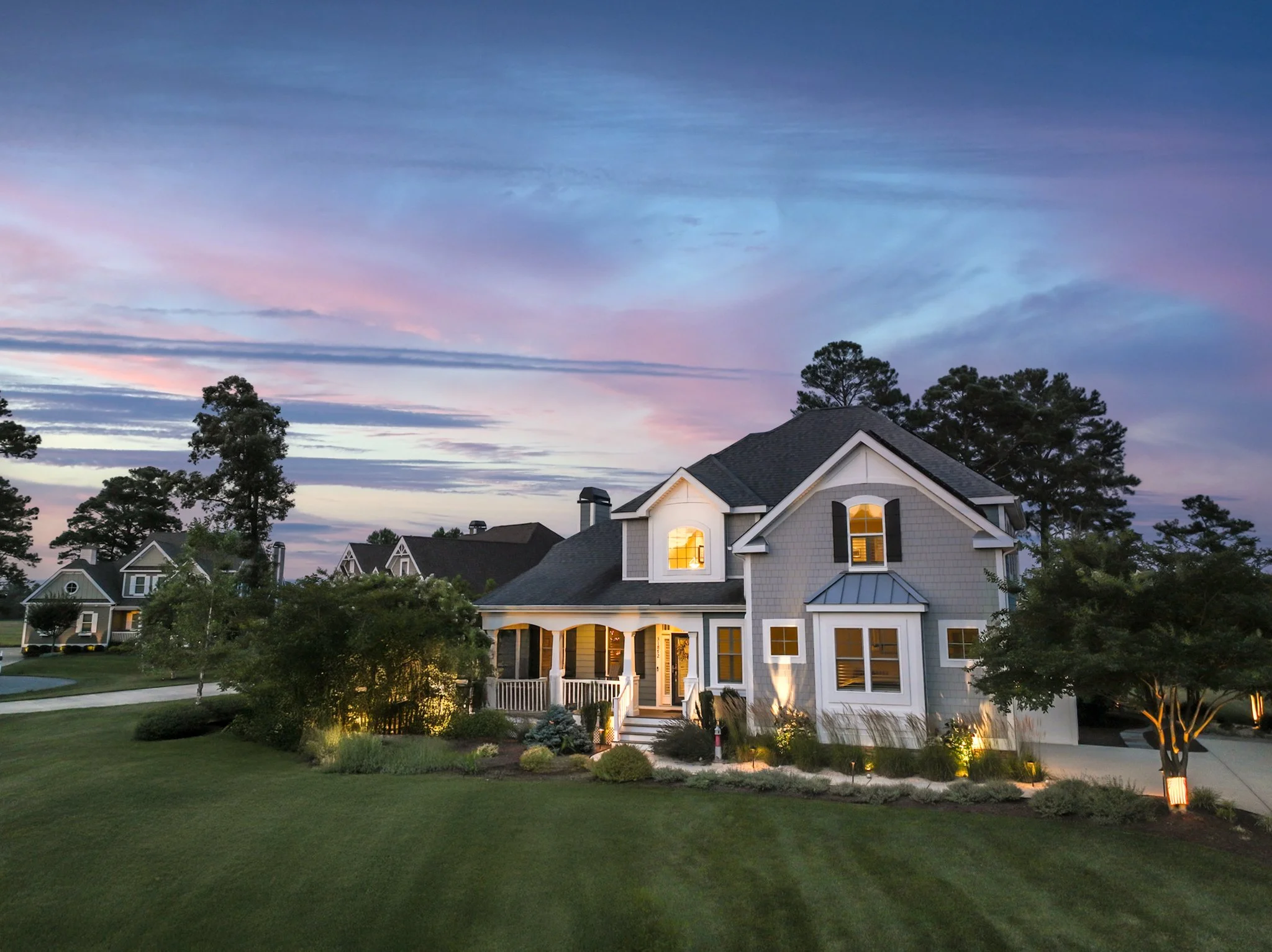 A suburban house with lit-up interior lights at dusk, surrounded by a well-manicured lawn and trees, with a cloudy sky showing hints of pink and purple.