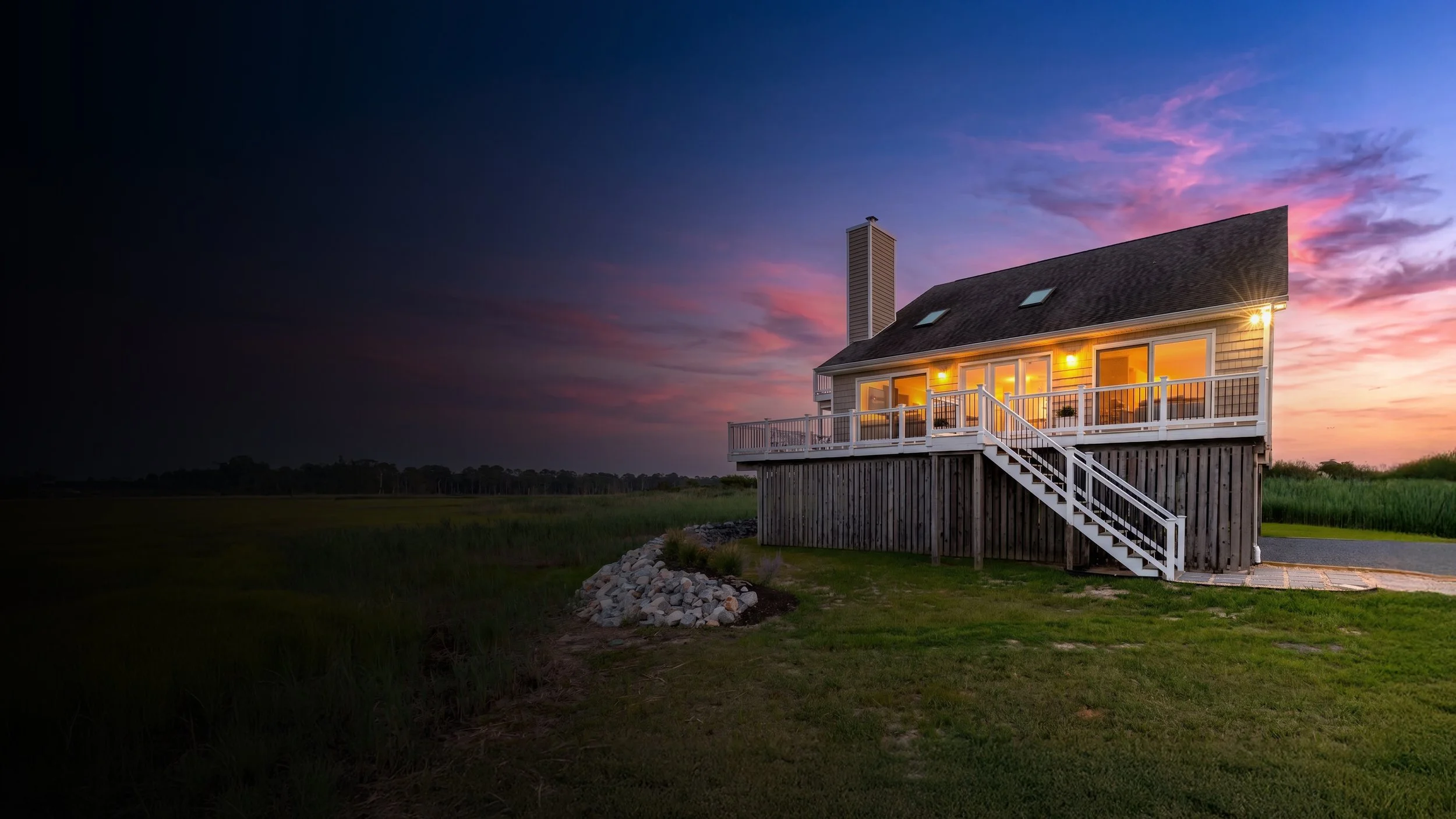 A house with large windows and a balcony in a rural area at sunset, with a colorful sky and surrounding grassy fields.