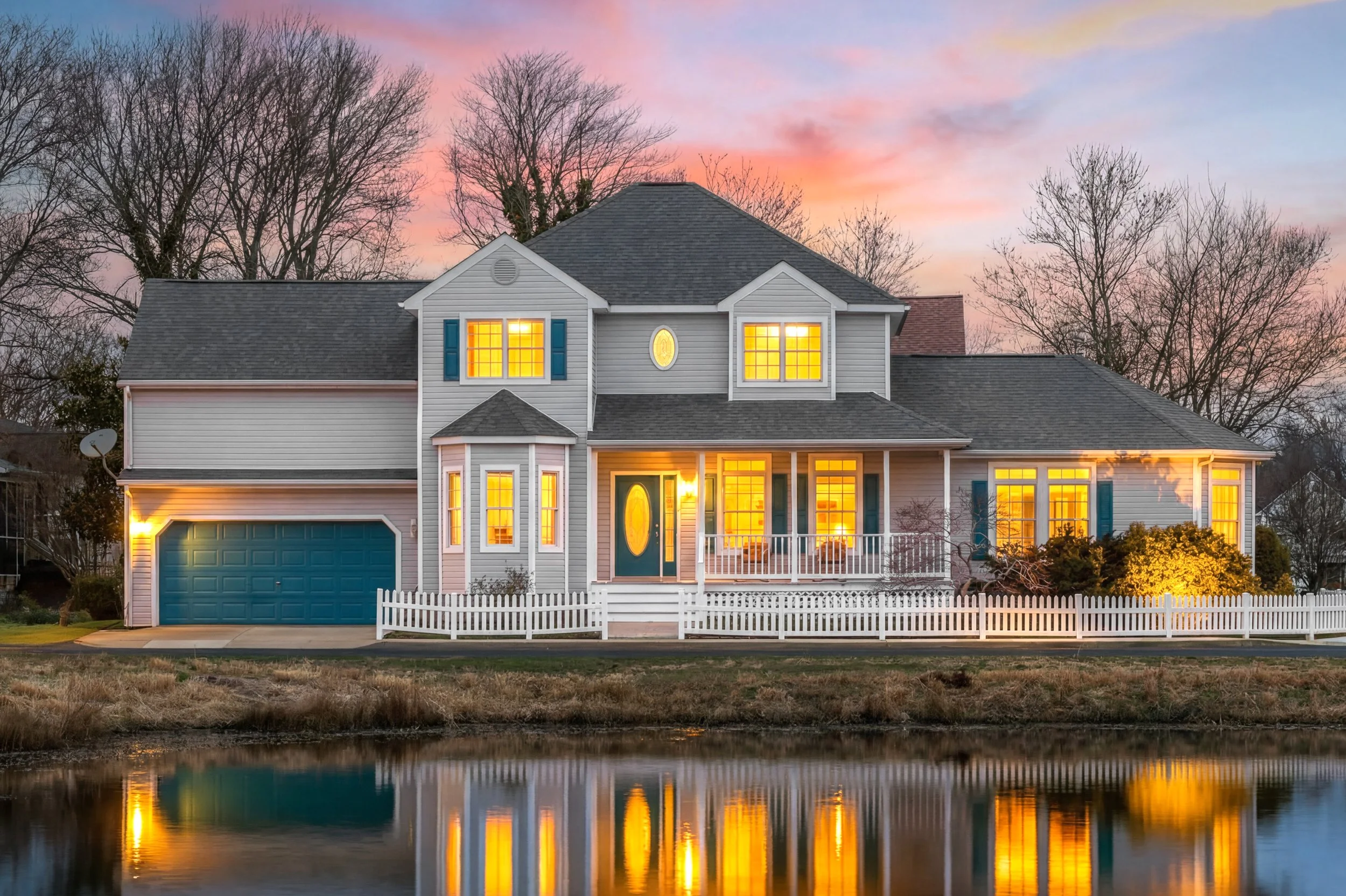 A two-story house at sunset with lights on, gray siding, a blue garage door, and a white picket fence, reflected in a nearby body of water.