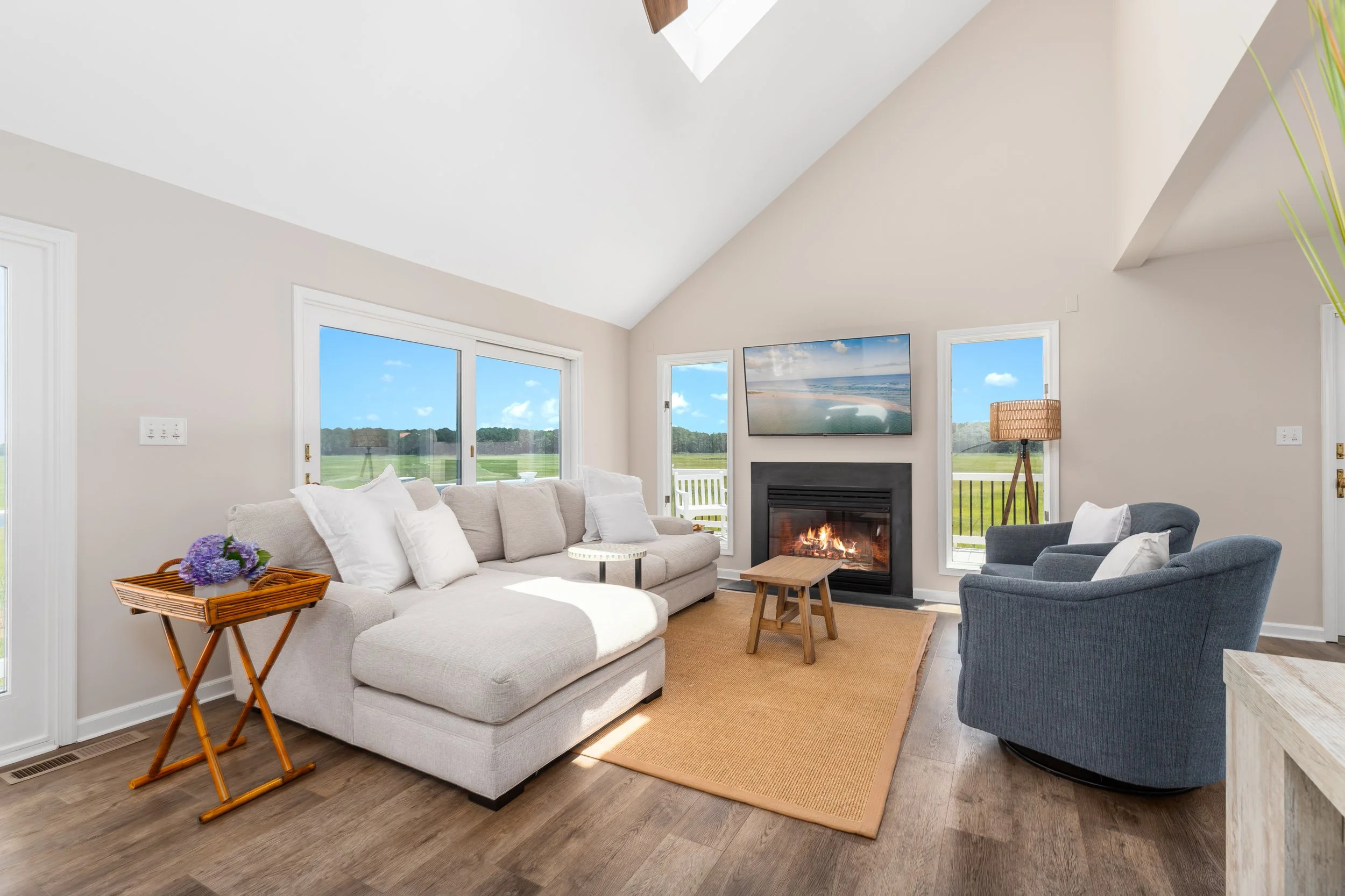 Living room with white walls, large windows showing a green outdoor landscape, a sofa with white pillows, a coffee table, a fireplace on the wall above a flat-screen TV, a floor lamp, a blue armchair, a small side table, and a wooden tray with purple flowers.