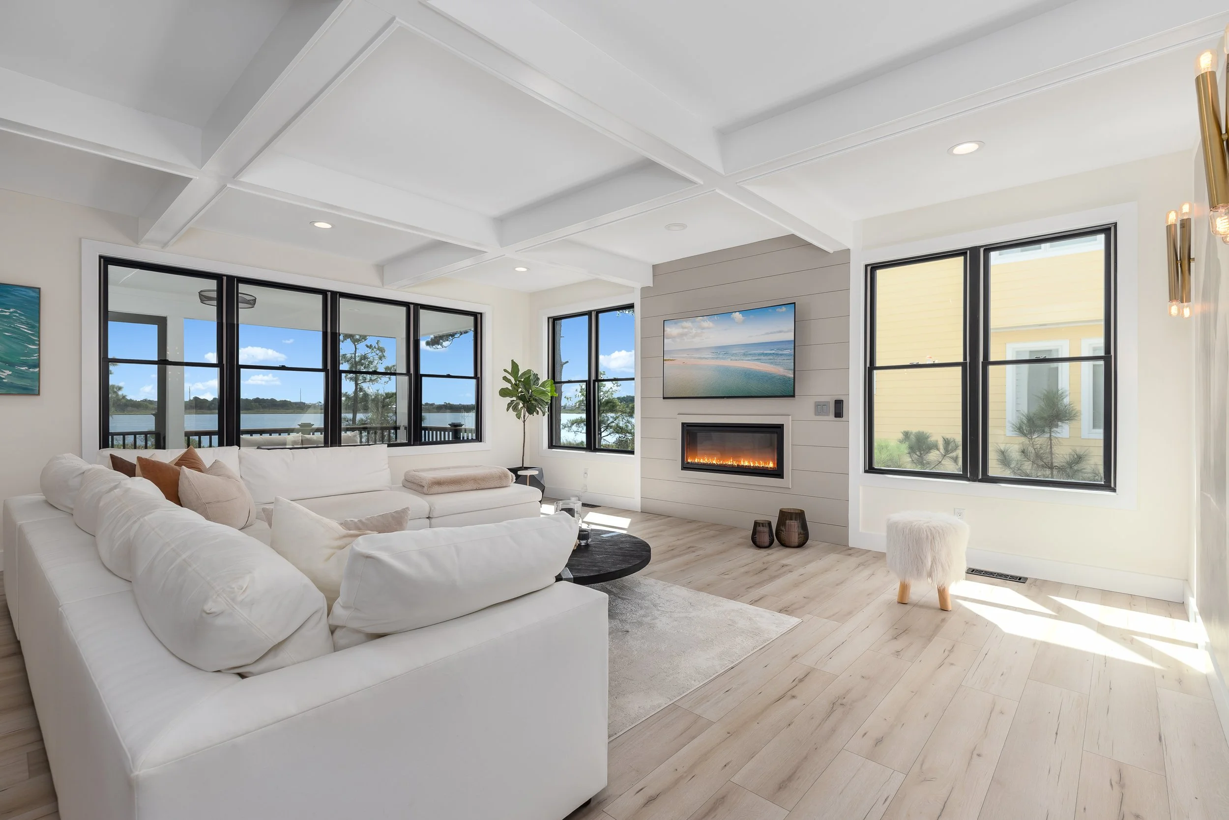 Real Estate Photography. Bright living room with white sofa, large windows with a water and landscape view, a fireplace with a TV above, a potted plant, and minimalistic decor.