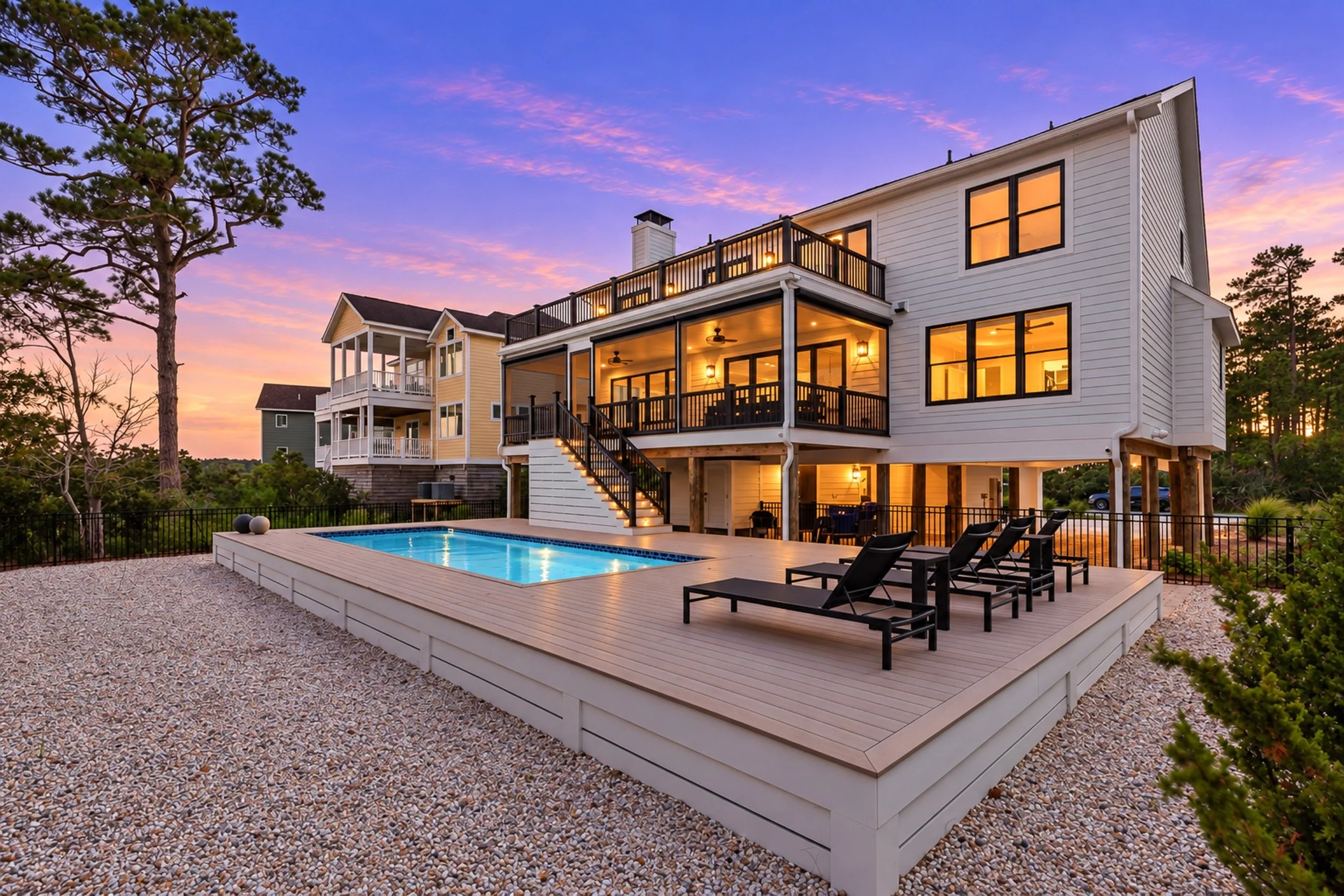 Real Estate Twilight Photography, a modern house with multiple large windows and a spacious balcony, illuminated from inside, during sunset. It has a backyard with a deck, black lounge chairs, a small swimming pool, and a fenced yard with trees.