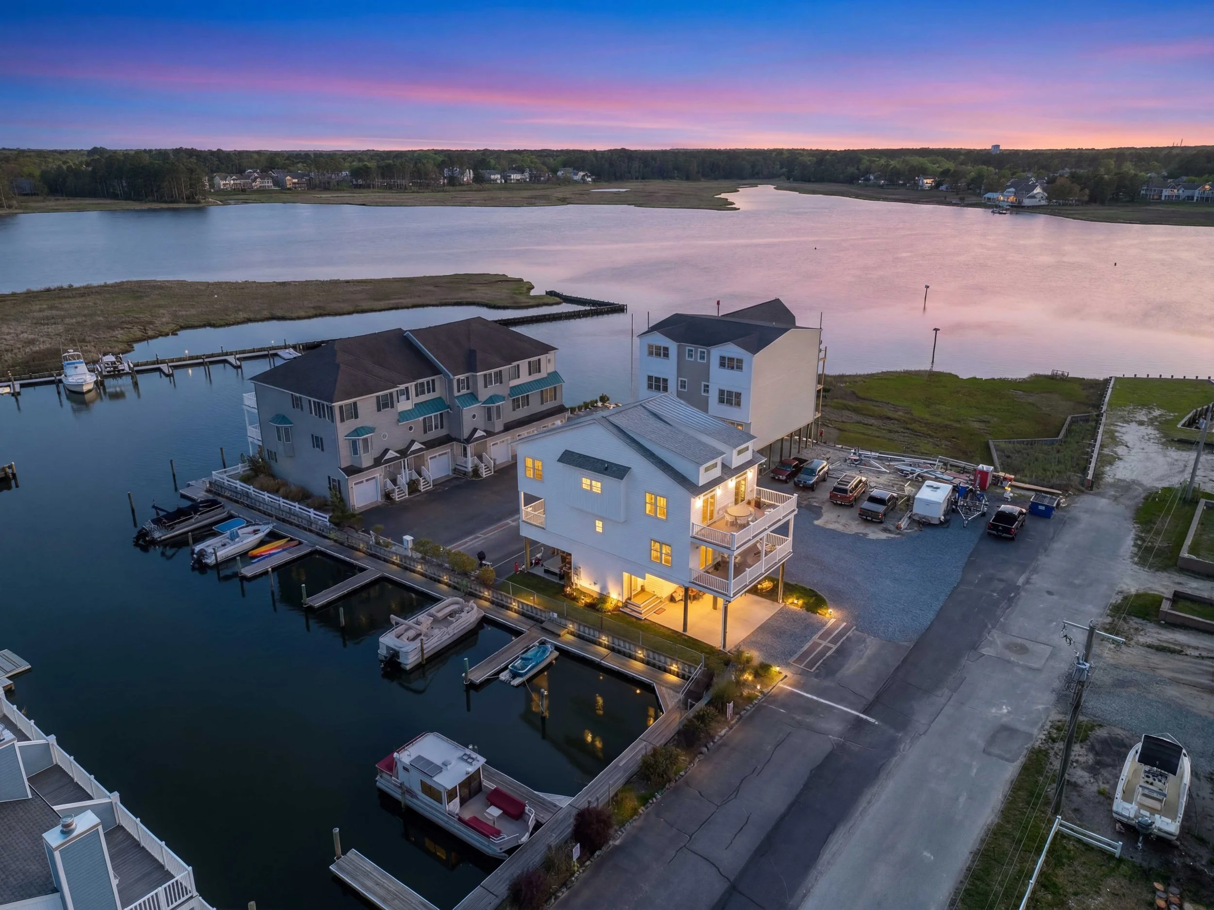 Aerial view of a coastal neighborhood at dusk with houses by the water and boats docked, pink and purple sky with clouds.