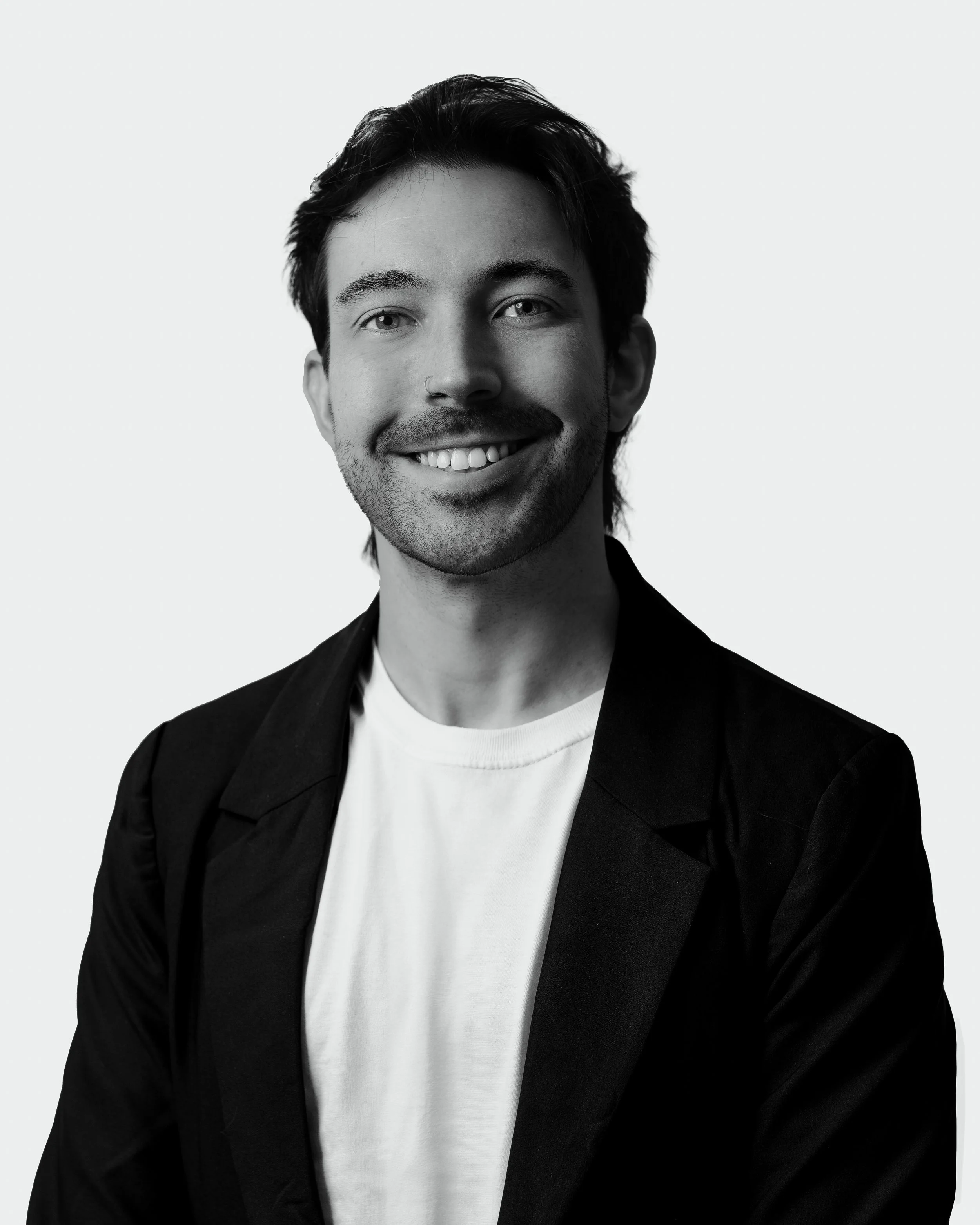 A black and white portrait of a young man with medium-length dark hair, a nose ring, and a short beard, smiling with his teeth visible, wearing a blazer over a white t-shirt, against a plain light background.
