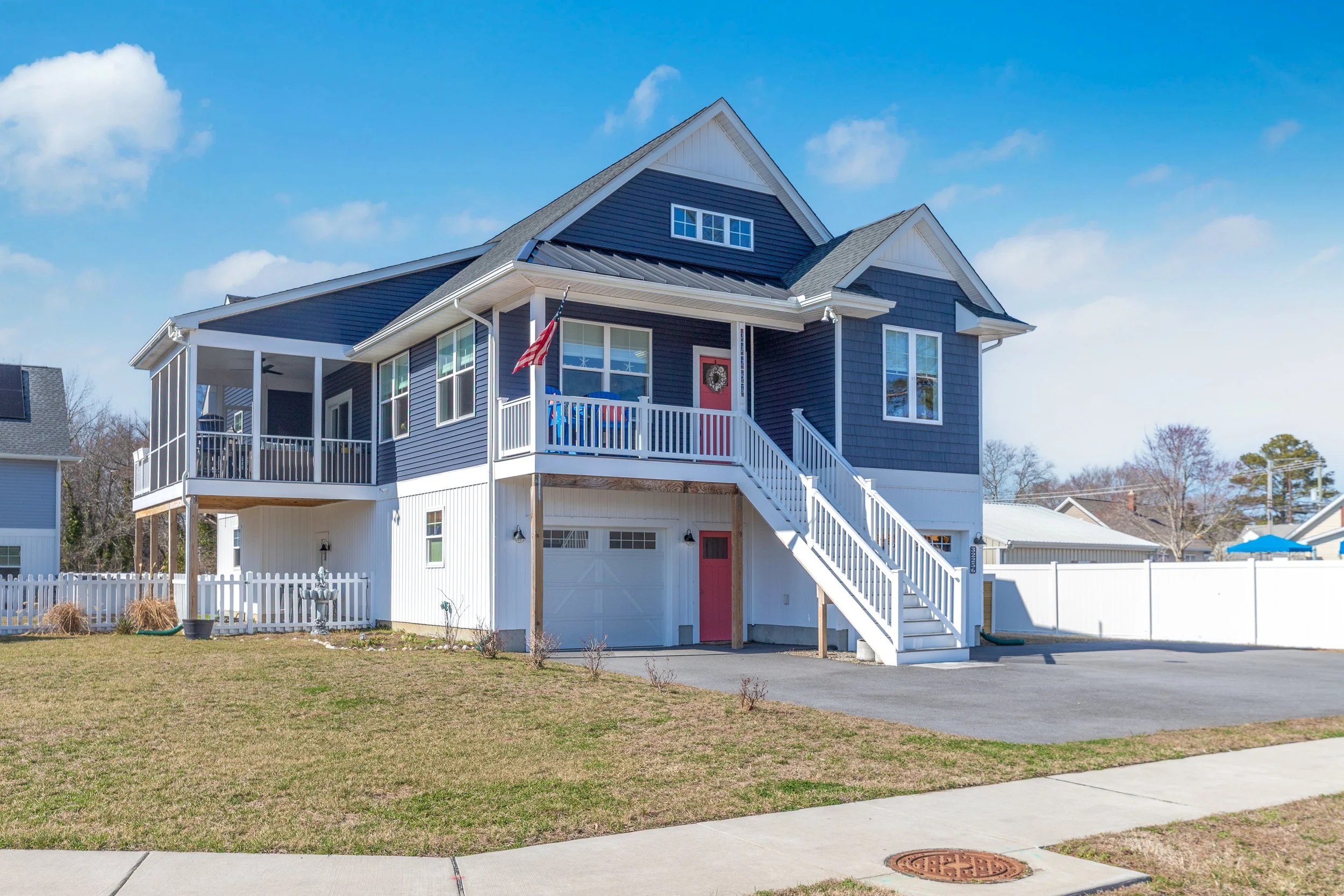 A modern two-story house with a dark blue exterior, white accents, a garage, and a front porch with stairs. An American flag and a wreath hang on the front door, and the yard has a small fence and mailbox.