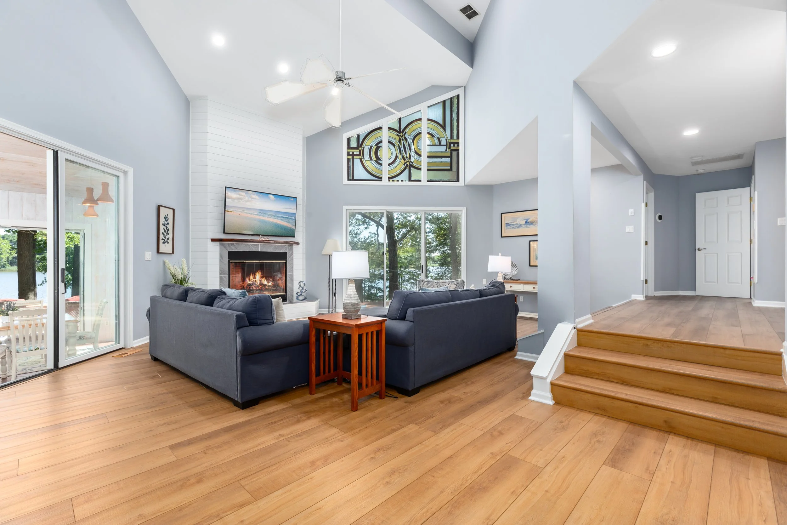 A spacious living room with light blue walls, wooden flooring, and large windows overlooking a lake. There is a fireplace with a TV above it, a dark-colored sofa set, and a wooden side table. Artwork and decorative items are on the walls, and a ceili