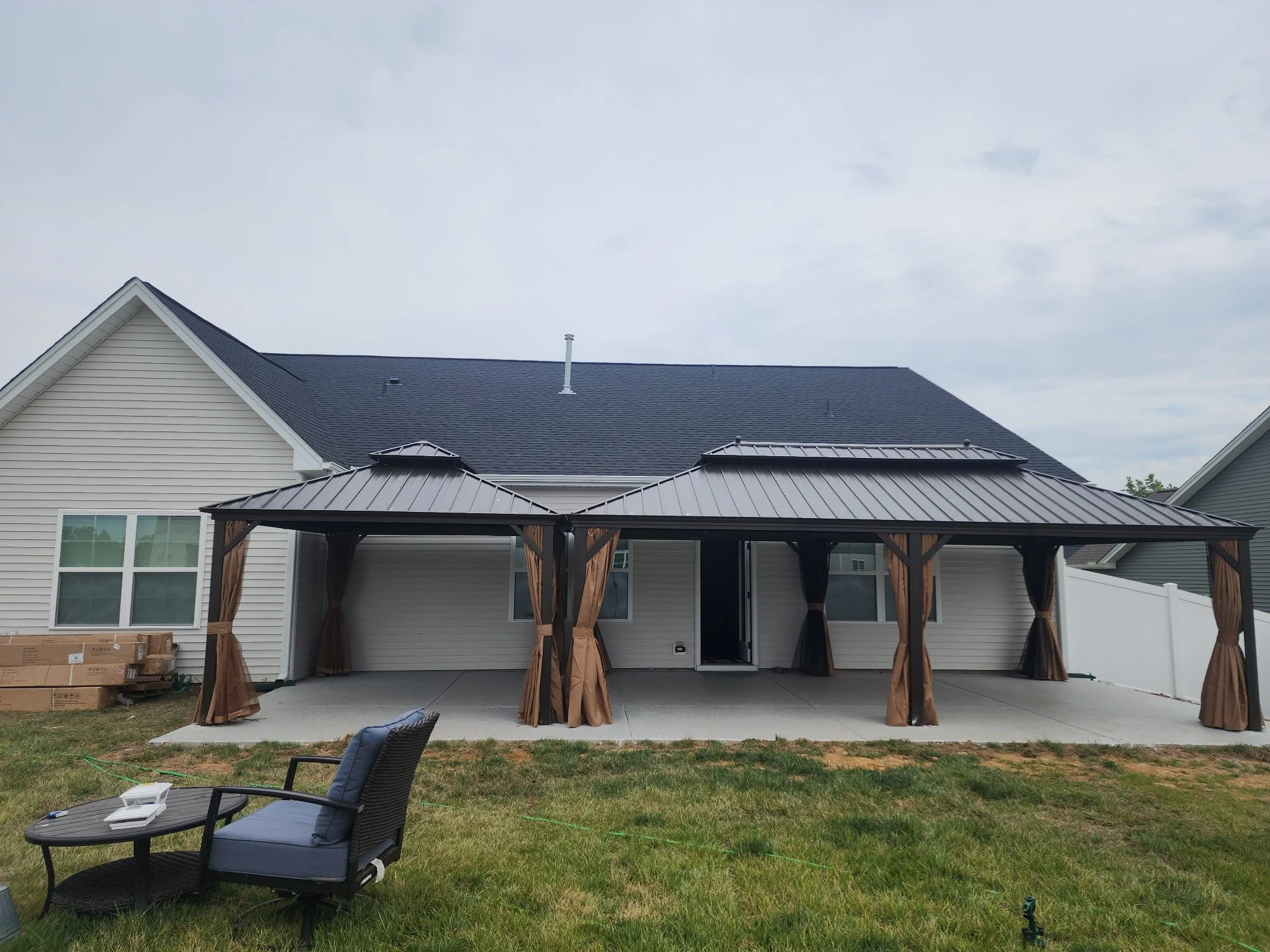 Backyard patio of a house with a metal gazebo and curtains, outdoor chair and table, and grass yard with a cardboard box and a few supplies.